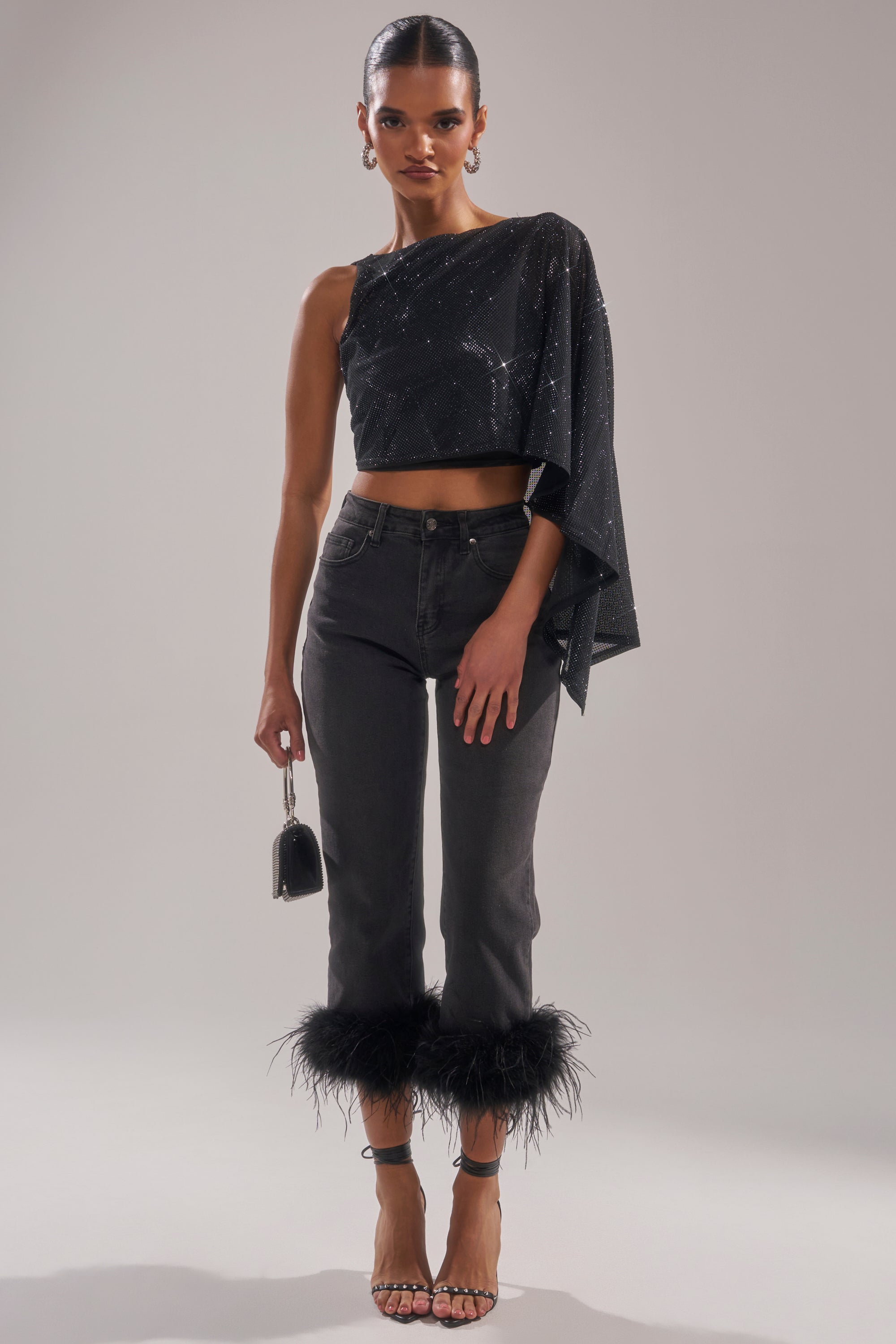 A woman poses in the TOO HOT FOR YOU RHINESTONE MESH TOP, high-waisted dark jeans with feather-trim hems, strappy heels, and a small black purse. Slicked-back hair and large hoop earrings complete her chic look. 7