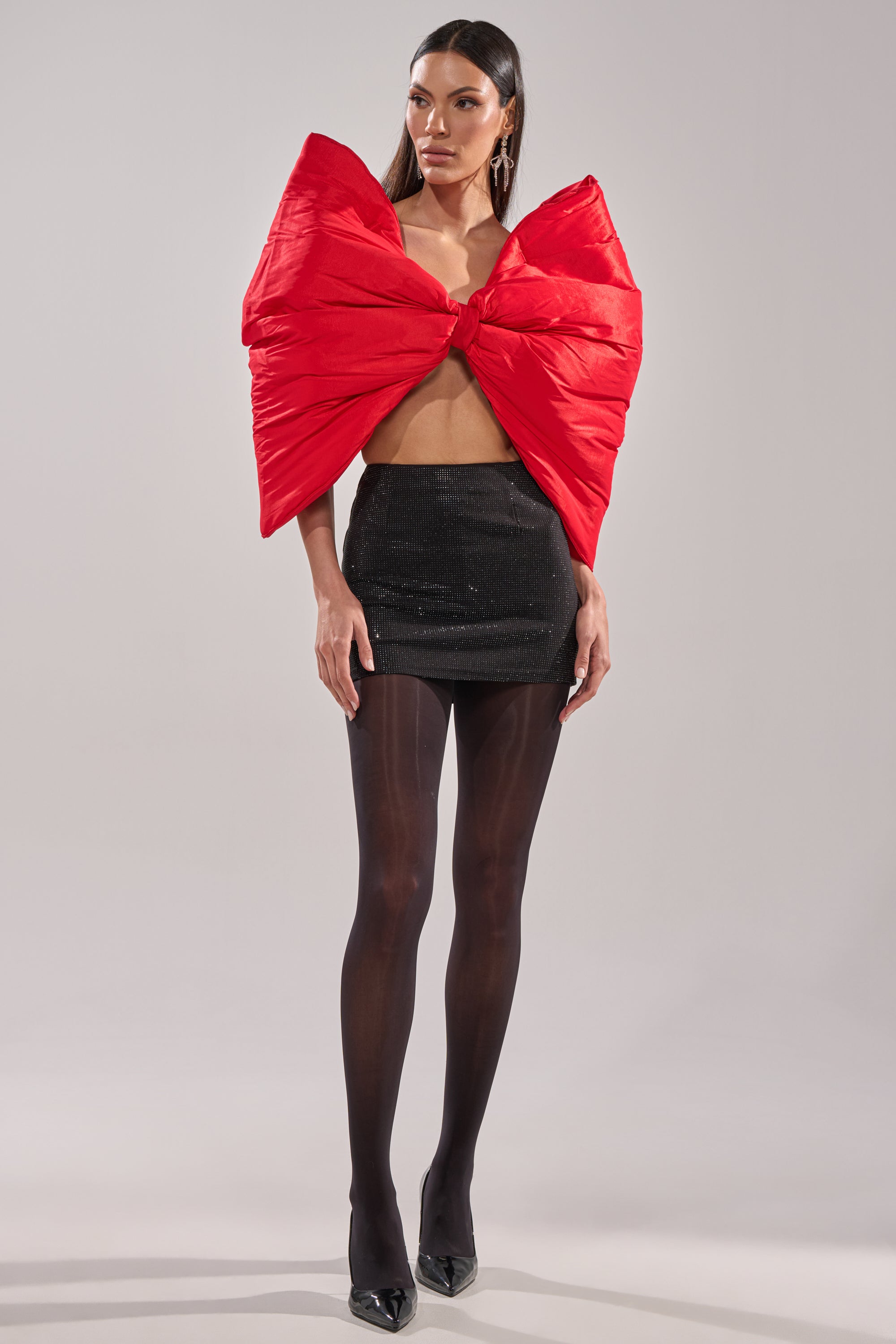 A woman stands against a plain light background wearing a bold red oversized bow-shaped top, the TOO HOT FOR YOU RHINESTONE MINI SKIRT in black, sheer black tights, and black pointed-toe heels. 5