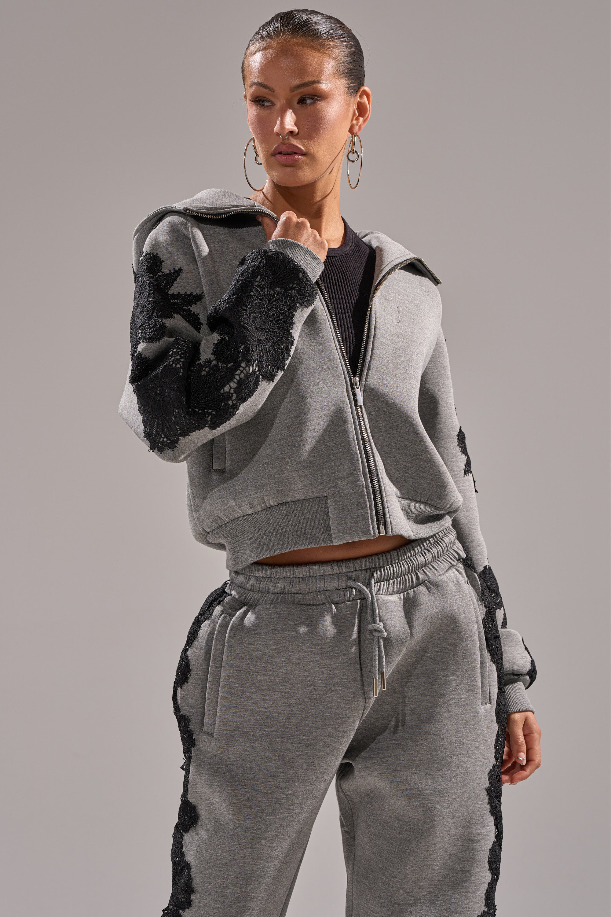 A woman poses with one hand on her hip, wearing the RAQUEL ZIP UP SWEATSHIRT in grey scuba material with black floral lace details and matching joggers, set against a plain background.
