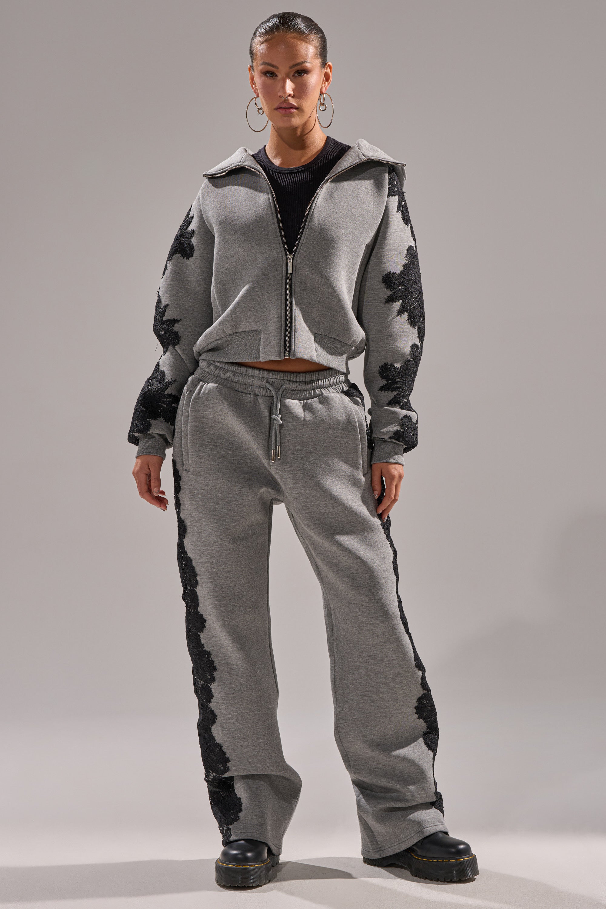 A woman wears a matching light gray RAQUEL ZIP UP SWEATSHIRT and jogger pants with black floral appliqués, a black top, hoop earrings, and black platform shoes, posing against a plain gray background.
