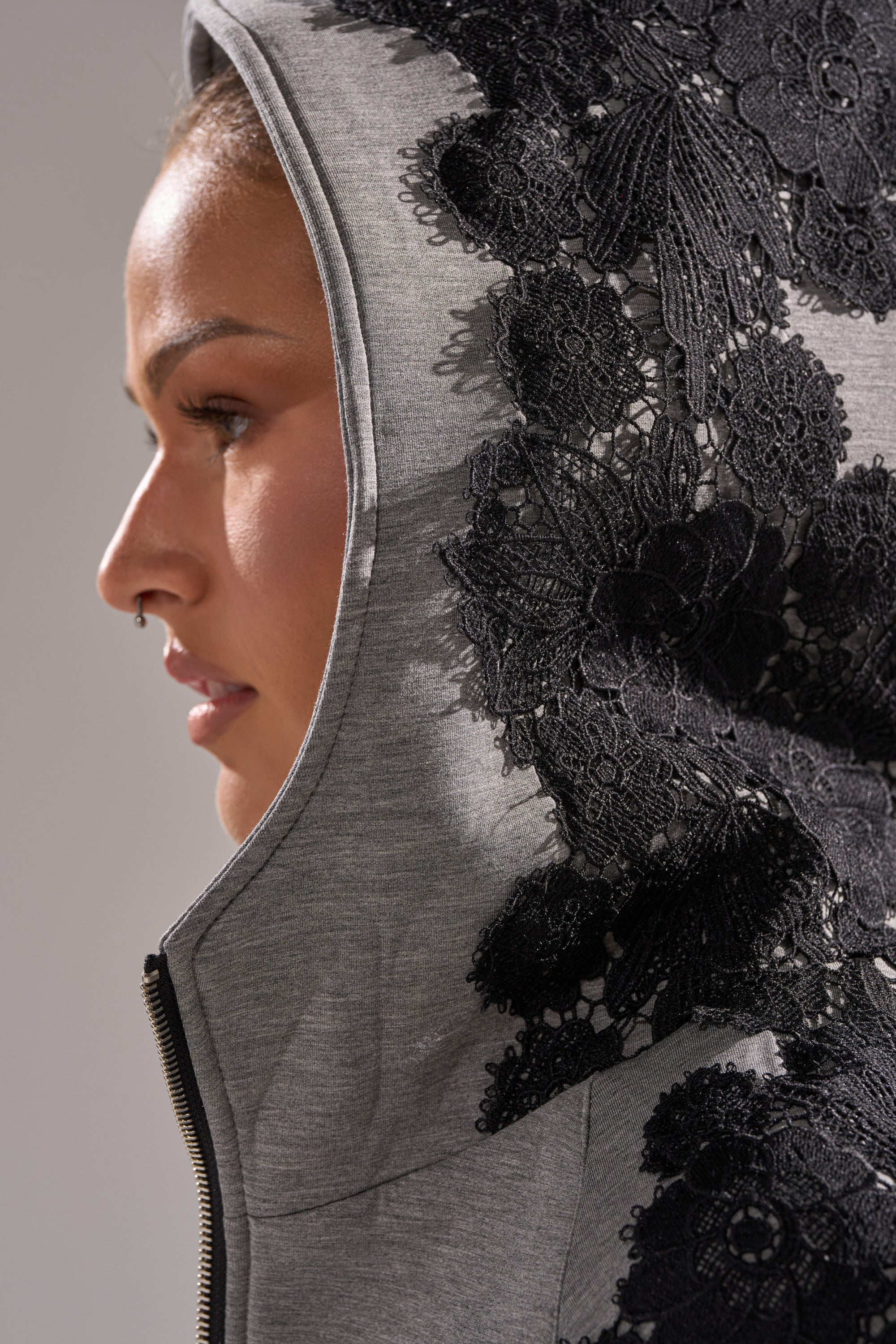 A side profile shows a person with a nose ring, looking left, wearing the RAQUEL ZIP UP SWEATSHIRT made from soft scuba fabric with detailed black floral lace appliqué.