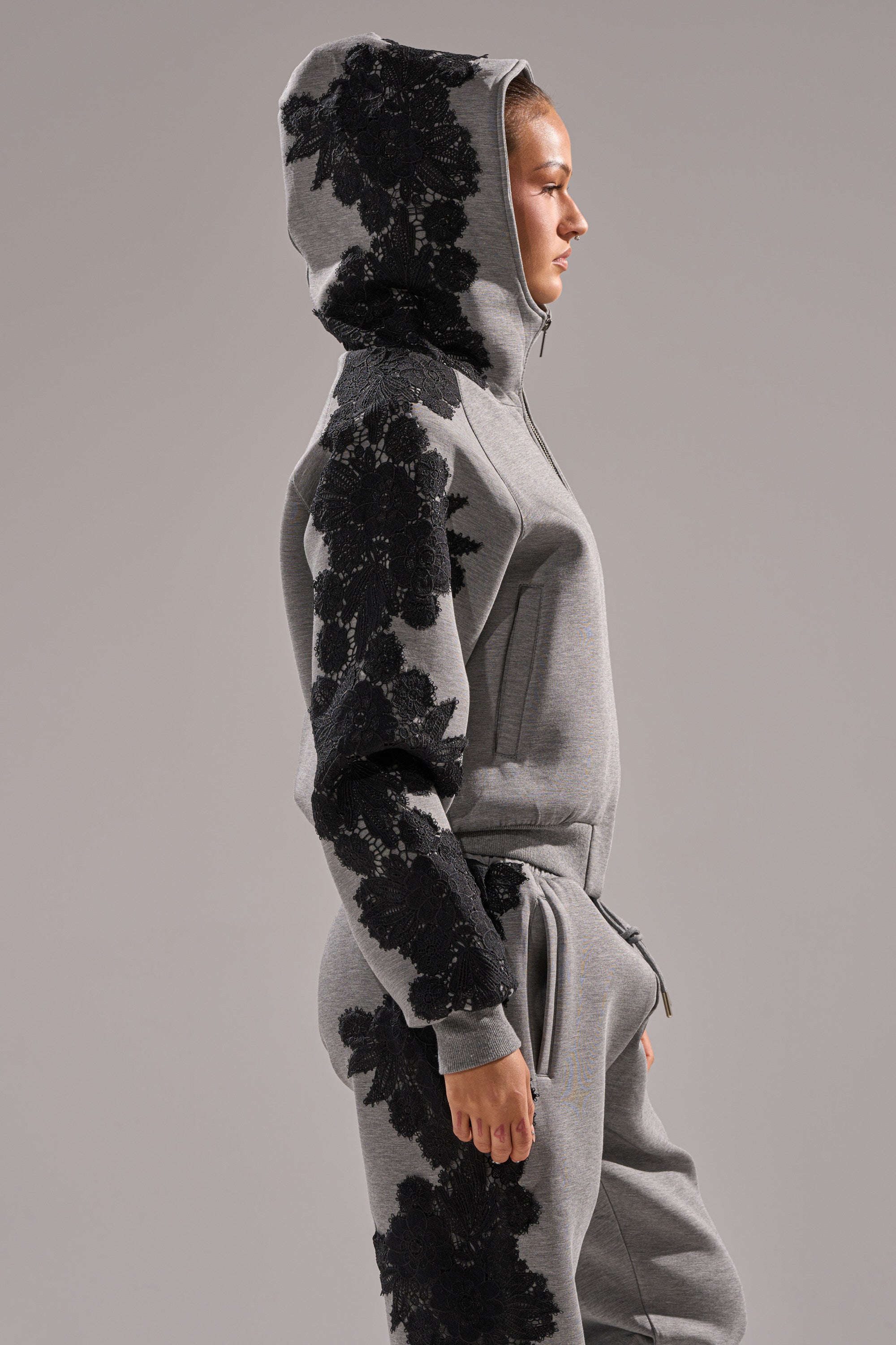 Someone stands in profile against a plain grey background, wearing the RAQUEL ZIP UP SWEATSHIRT in grey scuba fabric with bold black floral designs on the sleeve and side.