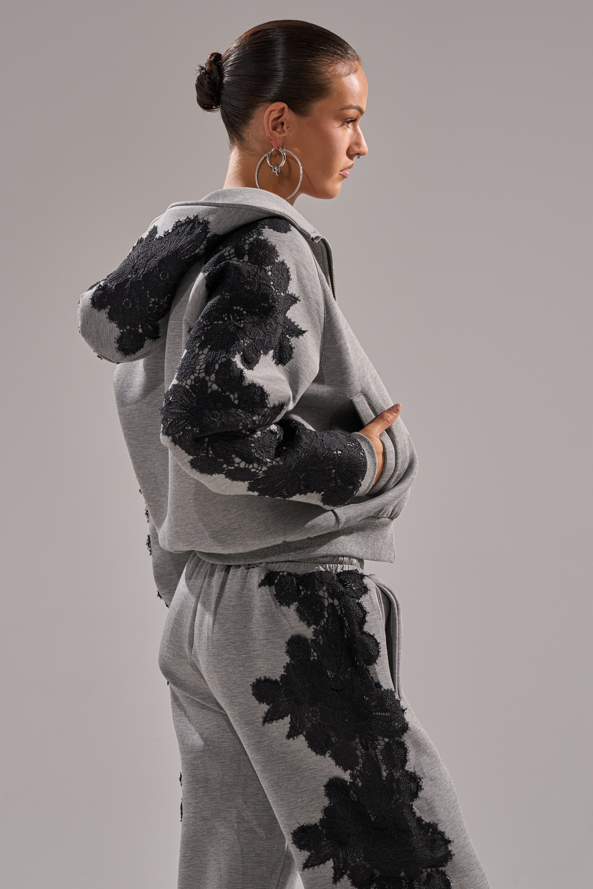 A woman with a bun models the RAQUEL ZIP UP SWEATSHIRT tracksuit in grey, featuring black floral lace appliqué on the scuba sleeves and legs. She poses in profile with one hand in her pocket against a neutral background.