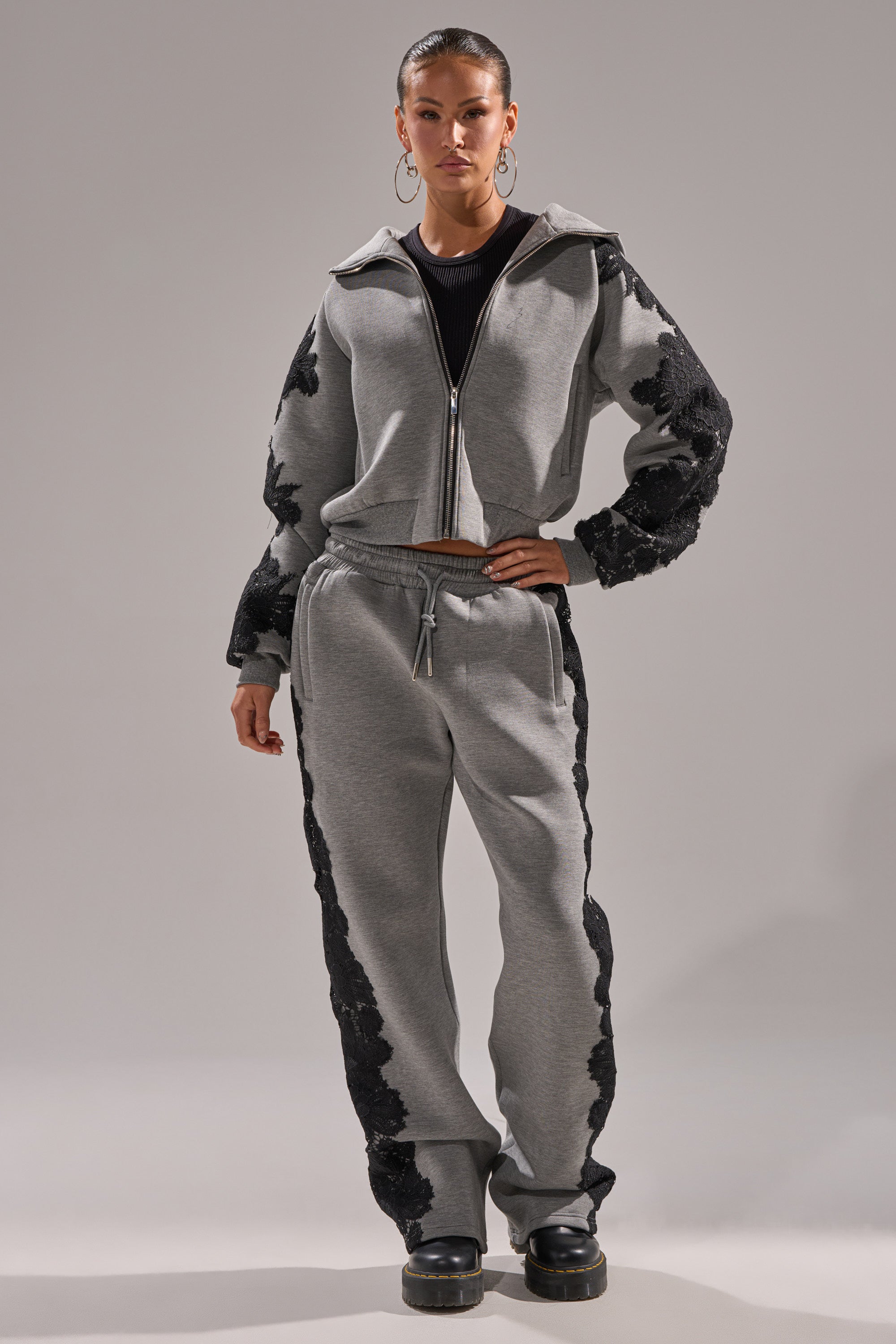 Wearing the RAQUEL ZIP UP SWEATSHIRT and matching gray scuba sweatpants with black floral lace on sleeves and legs, paired with black platform shoes, a person stands confidently against a plain background.