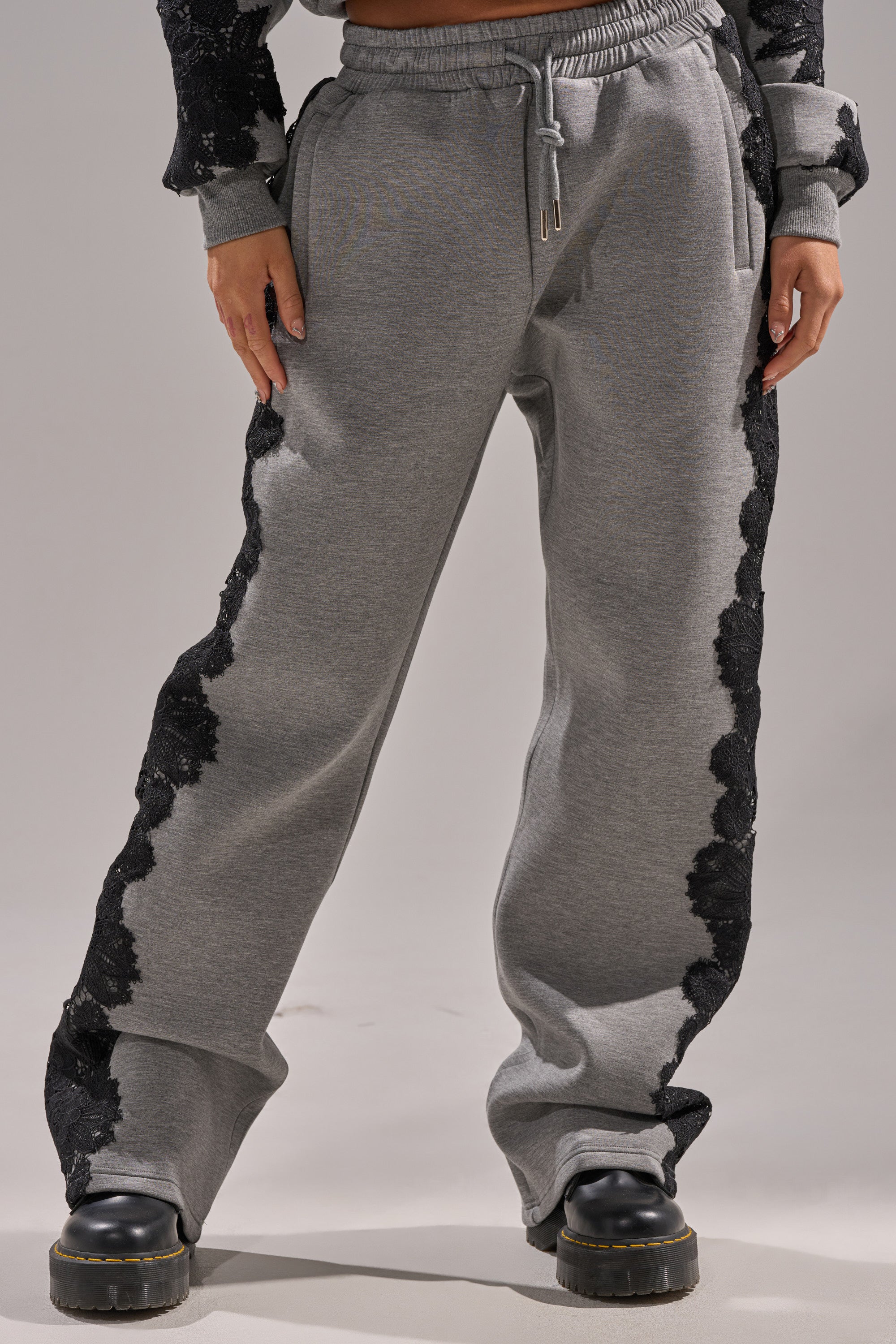 Model wears the RAQUEL STRAIGHT JOGGER PANT, a high-rise style with black floral lace along the sides, paired with chunky black shoes. The cropped photo shows hands in pockets and focuses on the joggers from shoulders down.