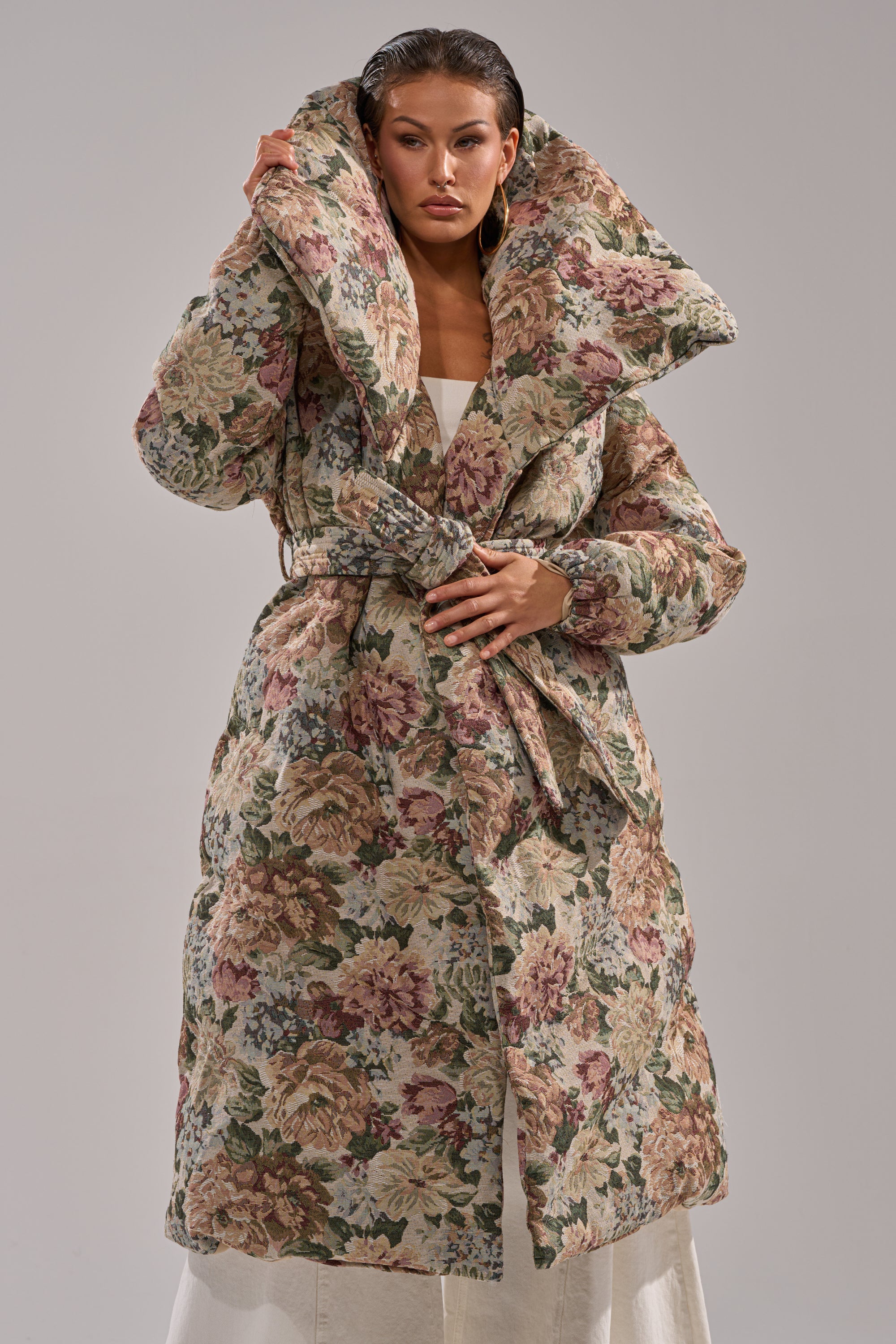 A woman wears the I'M OUTSIDE TAPESTRY PUFFER, a long floral jacquard coat with quilted insulation and an oversized collar, belted over a white outfit. She holds up the collar confidently, posing against a plain background.