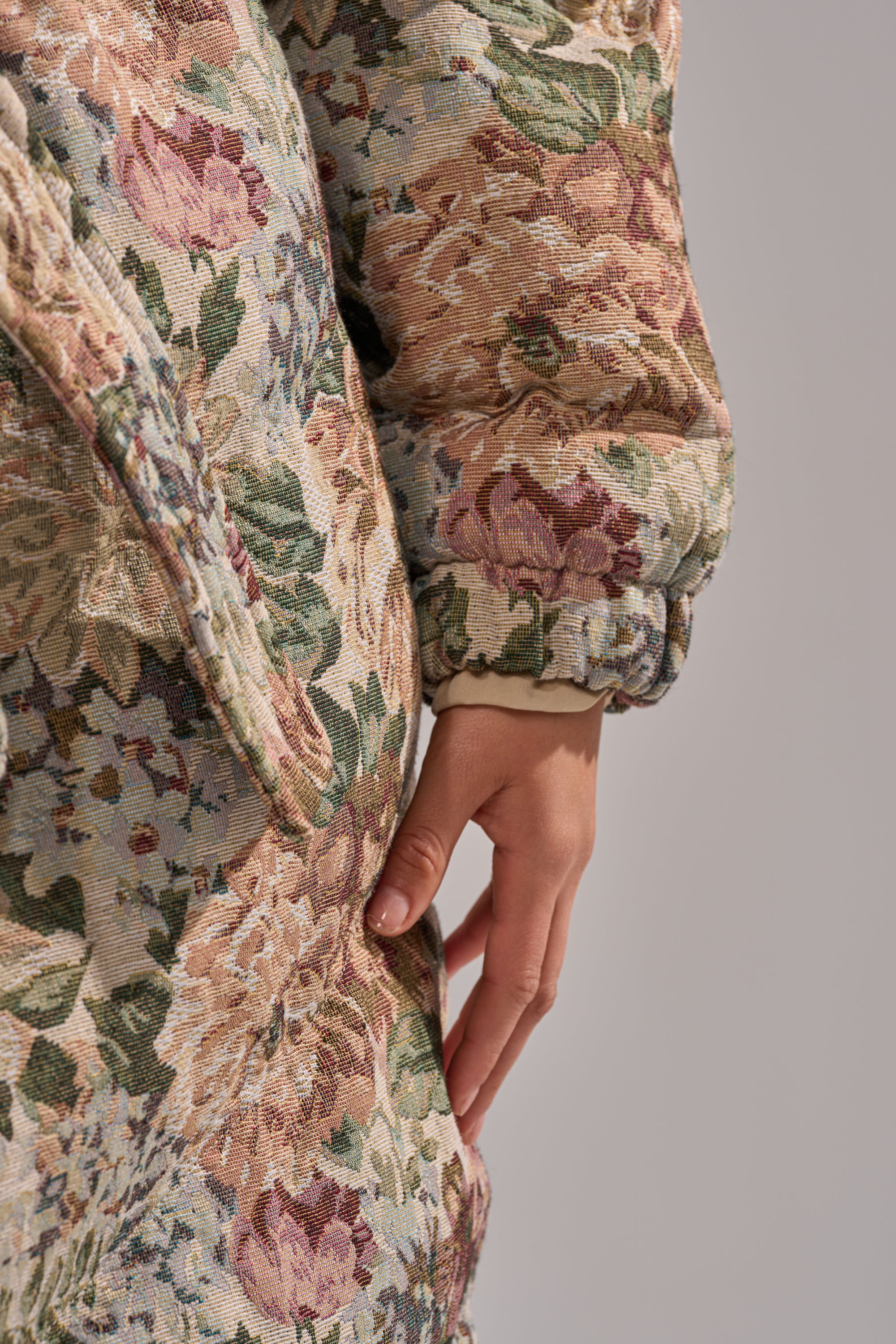 A close-up of a hand resting on the I'M OUTSIDE TAPESTRY PUFFER, showcasing textured fabric with pastel pink, green, and beige floral patterns against a plain soft gray background.