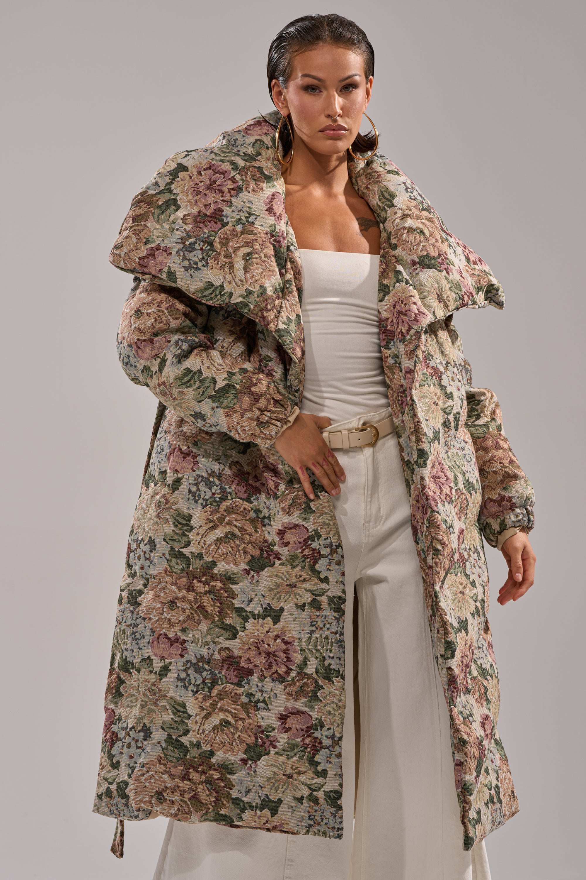 A woman stands confidently against a plain background in the I'M OUTSIDE TAPESTRY PUFFER, styled with a white top, wide-leg pants, slicked-back hair, and large hoop earrings.