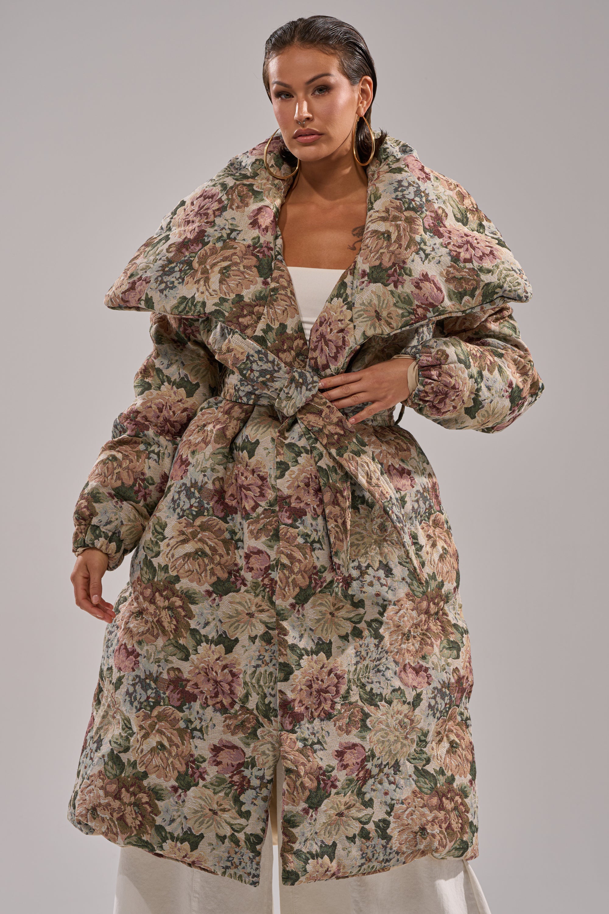 A woman poses confidently against a plain light gray background wearing the I'M OUTSIDE TAPESTRY PUFFER, a long jacquard puffer coat featuring floral patterns, a dramatic collar, and a matching belt.