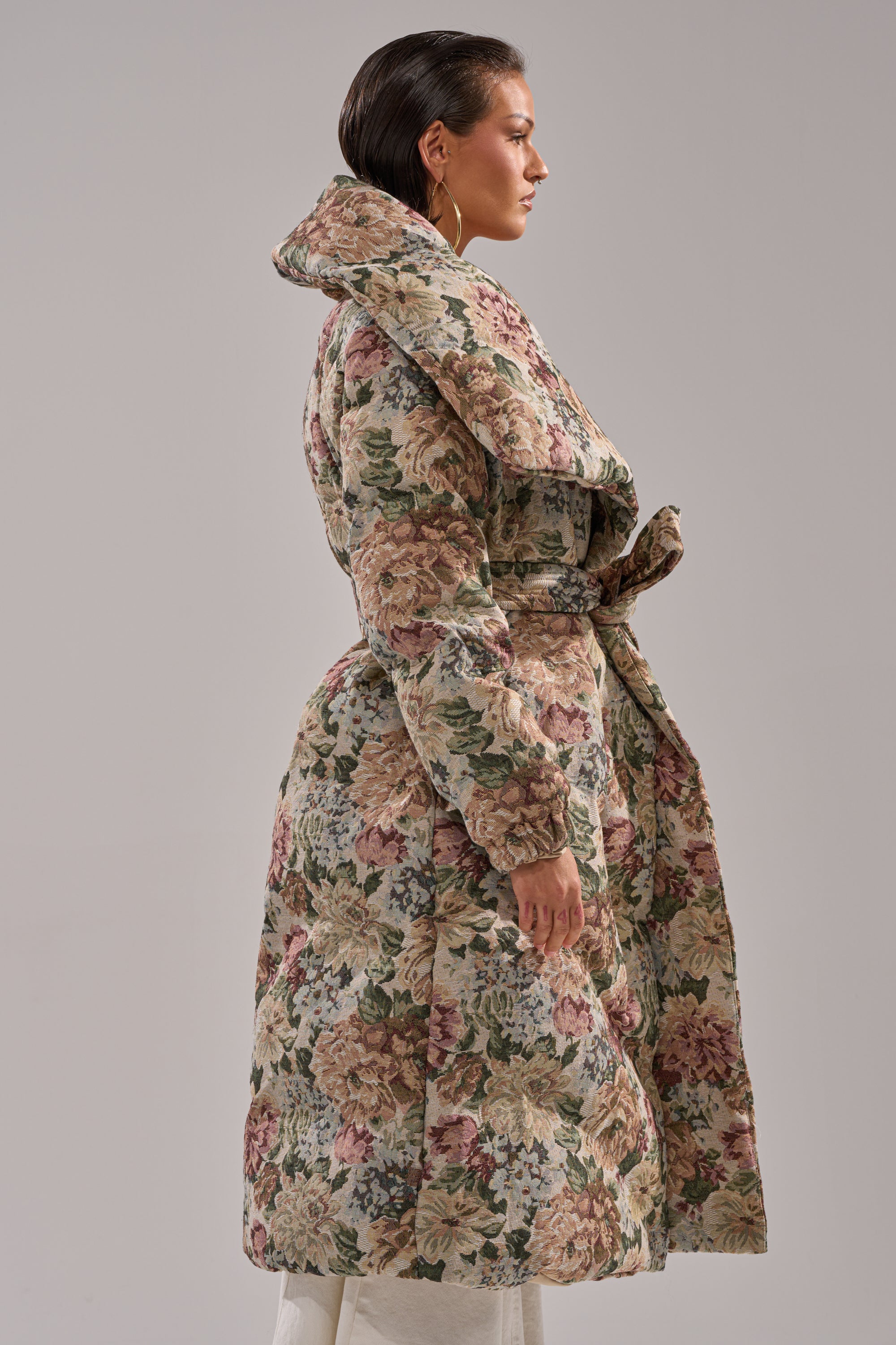 A woman in profile wears the I'M OUTSIDE TAPESTRY PUFFER, a full-length floral jacquard coat with a wide collar and belt, standing against a plain gray background.