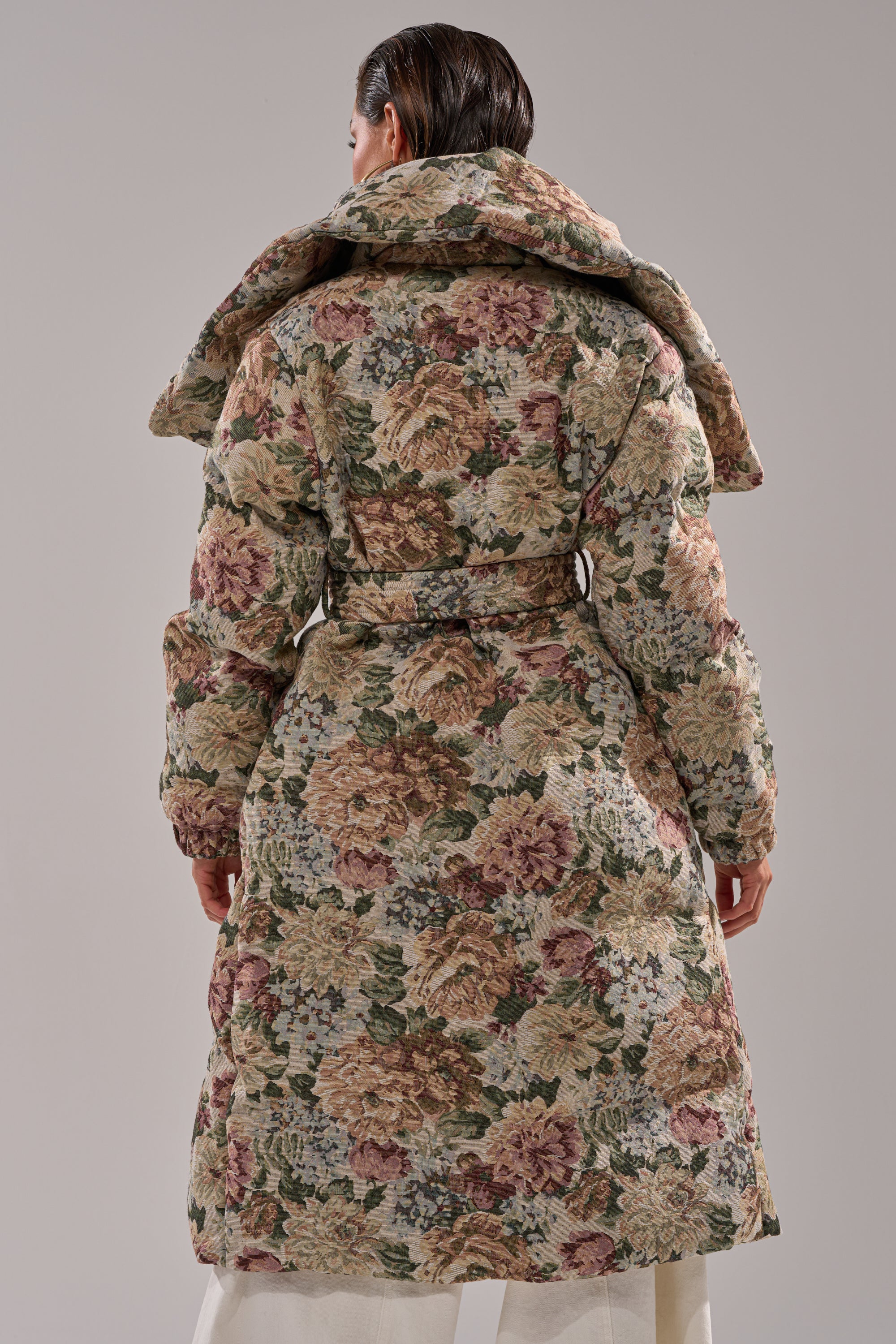 A person with wet, slicked-back hair wears the I'M OUTSIDE TAPESTRY PUFFER, a full-length jacquard quilted coat with an oversized collar, featuring bold pink, cream, and green floral patterns for a standout look.