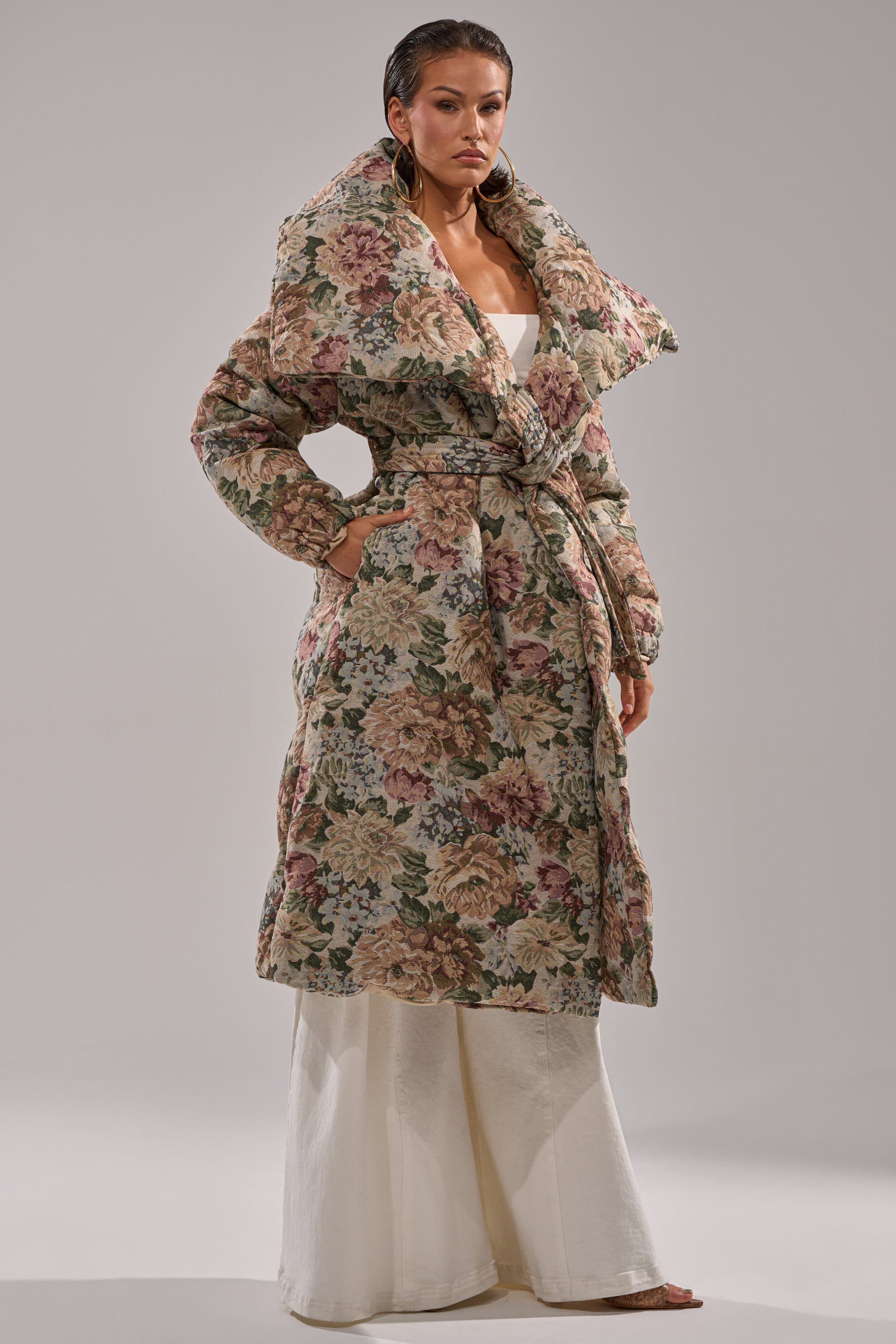 A woman wears the I'M OUTSIDE TAPESTRY PUFFER, a bold floral jacquard coat with an exaggerated collar, over a cream-colored dress. She gazes confidently at the camera against a plain background.