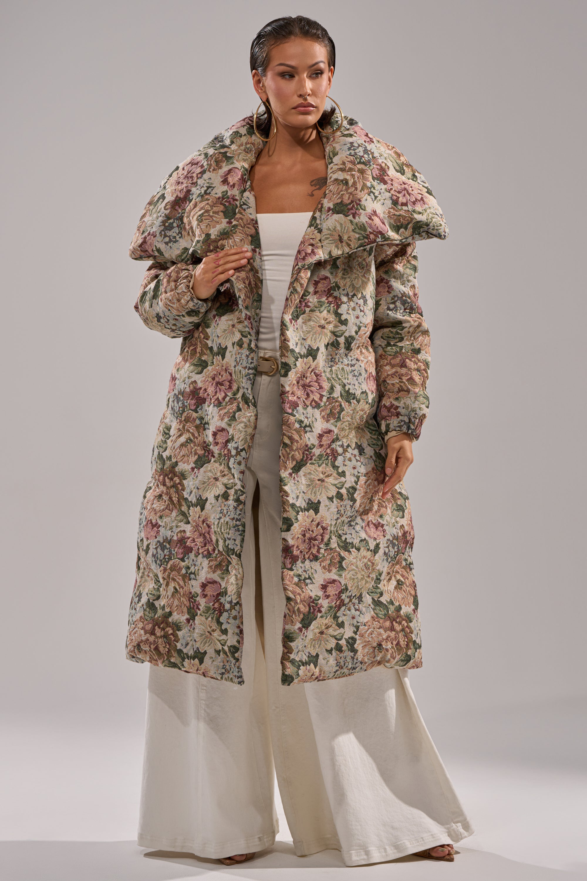 A woman stands confidently against a neutral background, wearing the I'M OUTSIDE TAPESTRY PUFFER over a white top and cream-colored wide-leg pants.