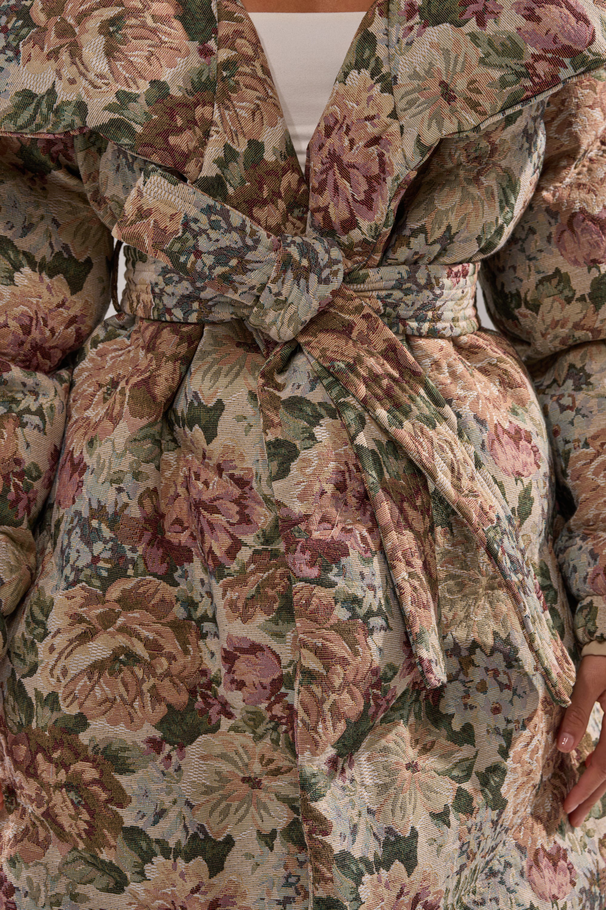 A close-up of a person wearing the I'M OUTSIDE TAPESTRY PUFFER, a full-length floral jacquard coat with a wide shawl collar and matching fabric belt. The muted pastel flower pattern gives it an elegant, tapestry-inspired style.