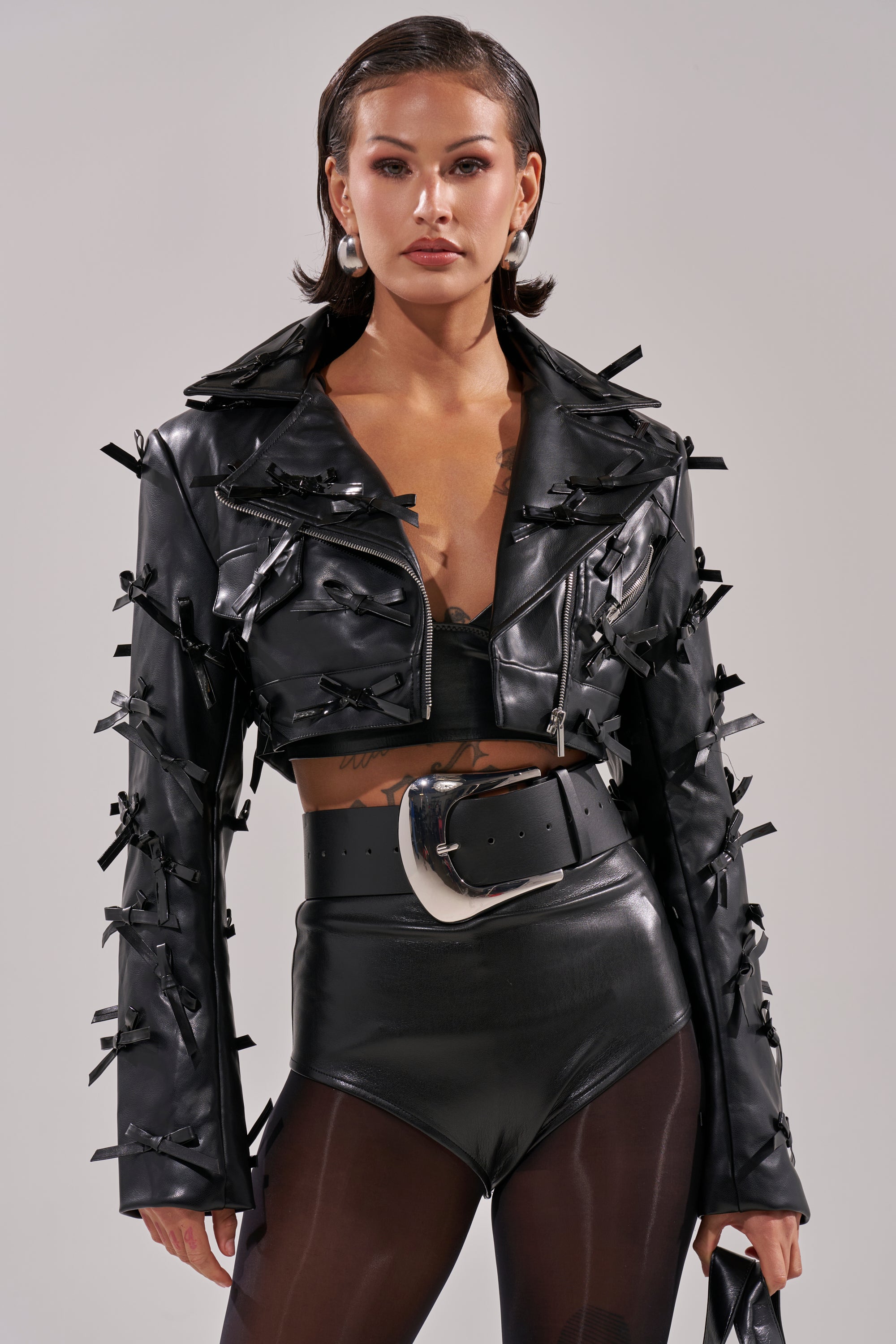 A woman wears the TELMO BOW MOTO cropped jacket with bow appliques, paired with black high-waisted shorts featuring a bold belt buckle and sheer black tights, standing against a plain background. 1