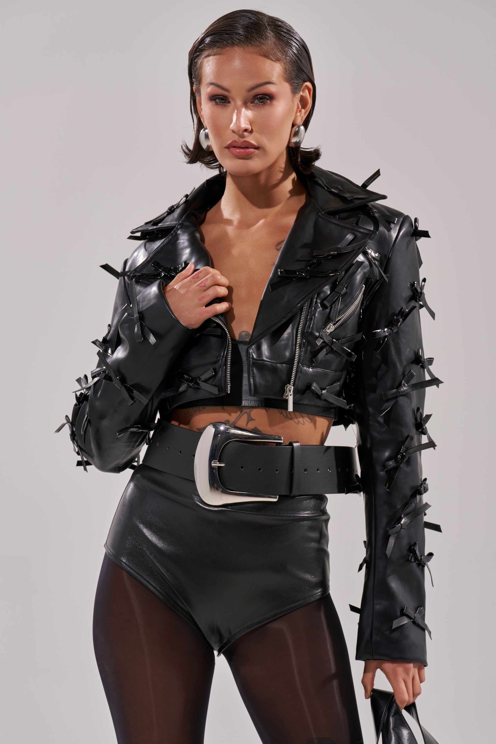 Wearing the TELMO BOW MOTO black cropped jacket with multiple bows, high-waisted black shorts, sheer tights, and a wide silver-buckled belt, the person stands against a plain background. 3