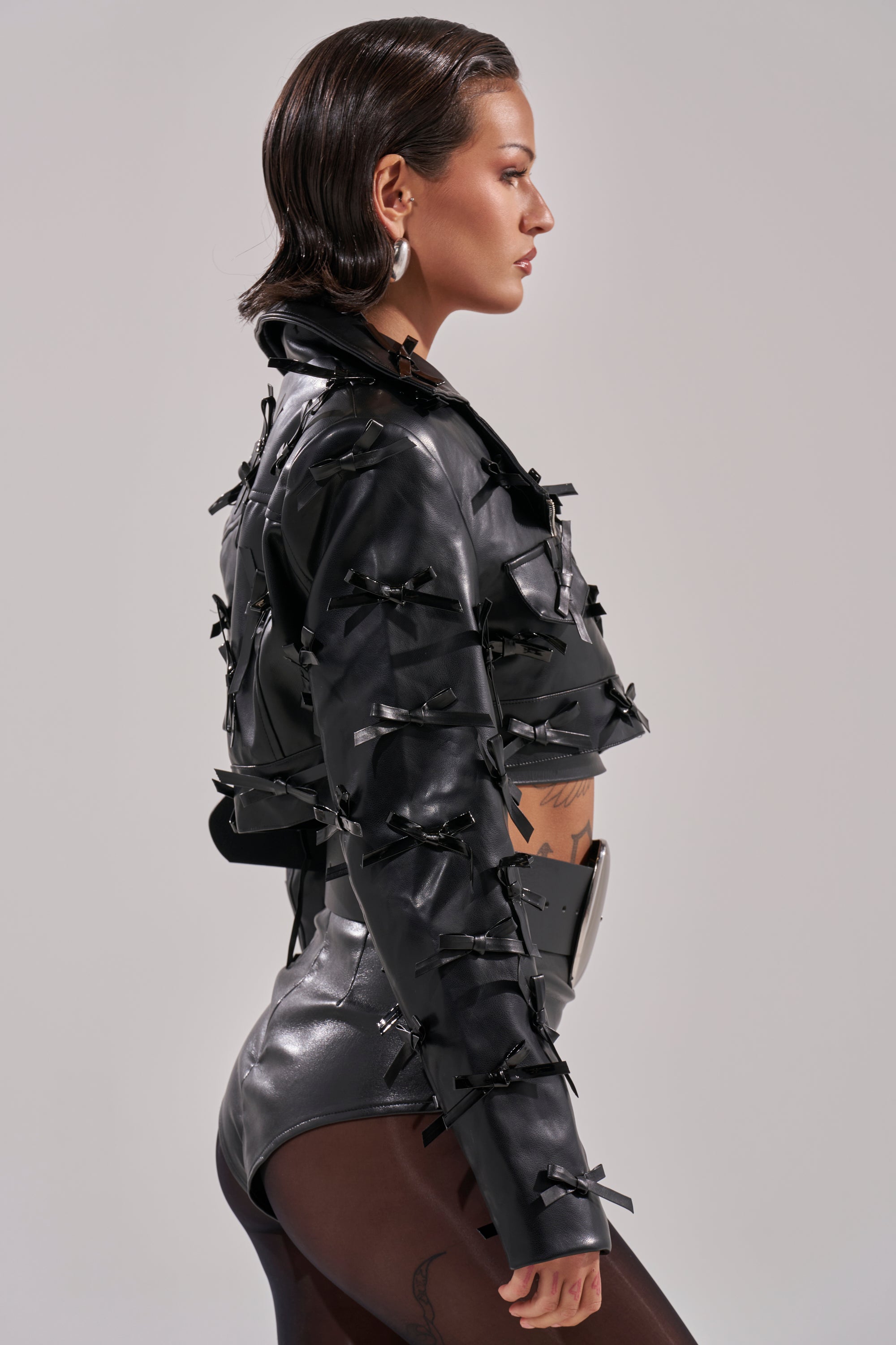 A woman in the TELMO BOW MOTO cropped jacket, high-waisted black bottoms, and sheer tights stands in profile against a neutral background with slicked-back hair and bold earrings. 5