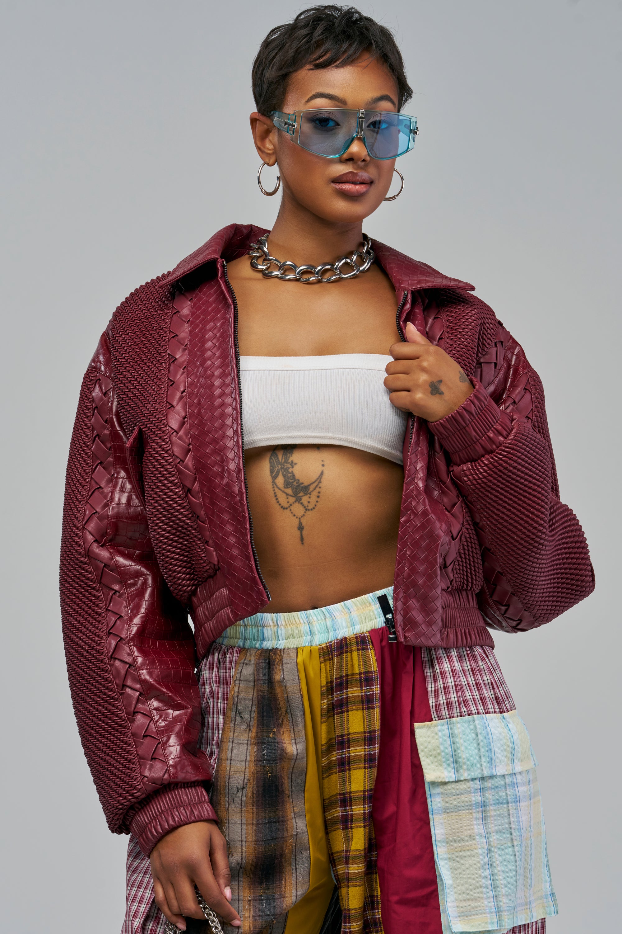 A person wears a KEEP IT LOWKEY PATCHWORK BOMBER IN BURGUNDY over a white bandeau top and multi-colored plaid patchwork pants, accessorized with large blue-tinted sunglasses, a chunky silver chain necklace, and fall styling.