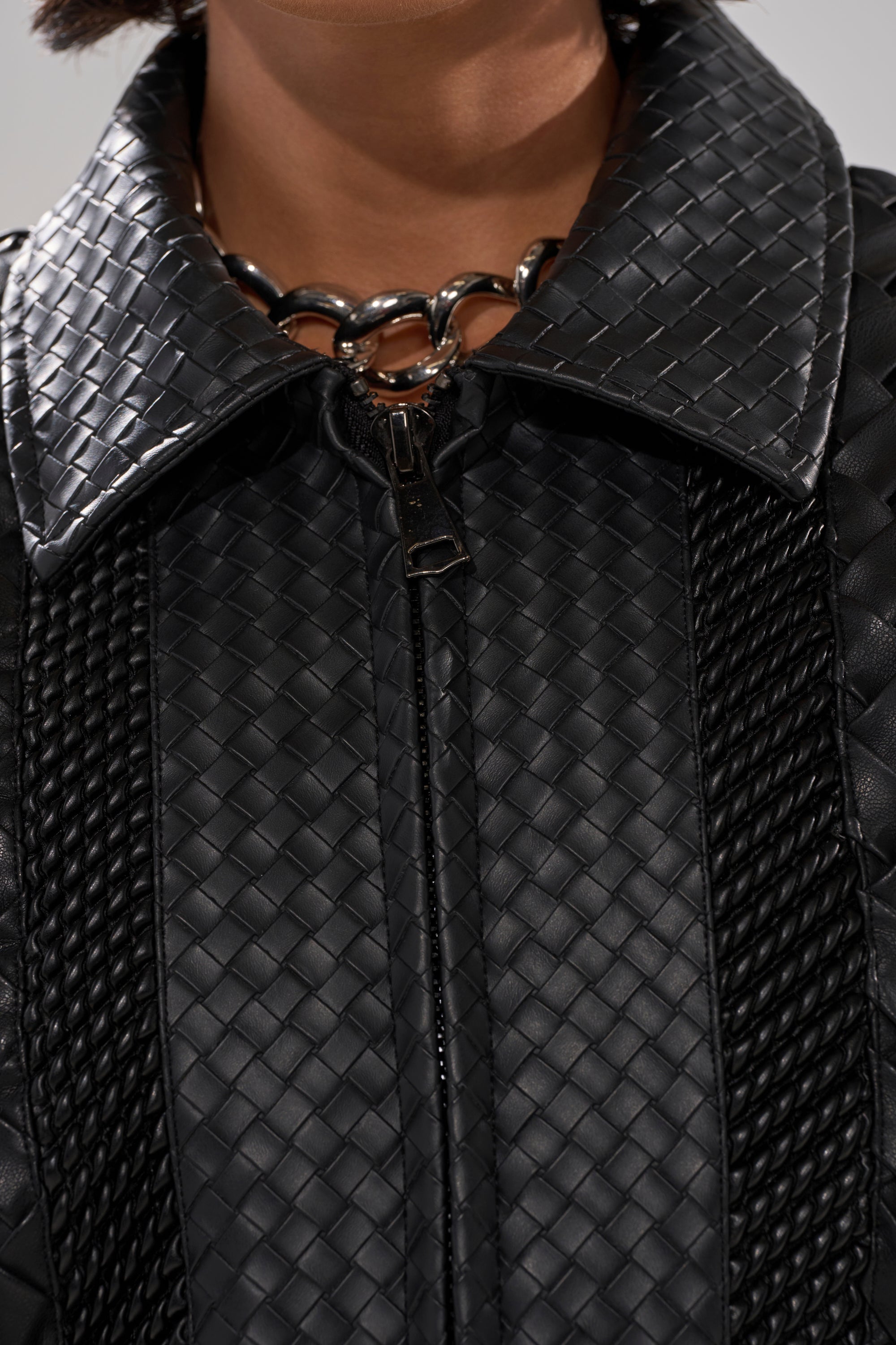 Close-up of the KEEP IT LOWKEY PATCHWORK BOMBER in black woven leather with a large collar, zipper closure, and textured details. A chunky silver chain necklace is partially visible at the neckline.