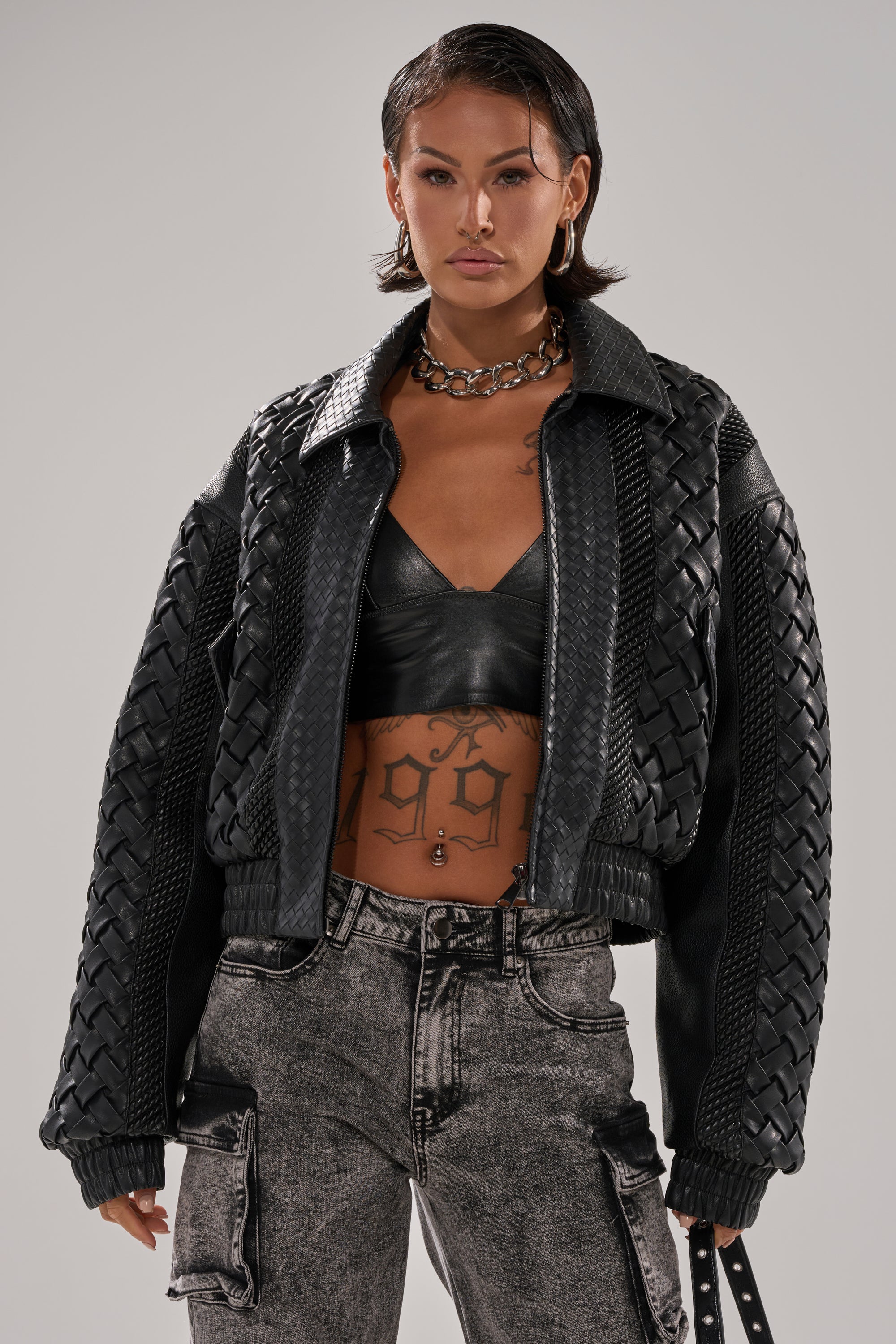 Wearing the KEEP IT LOWKEY PATCHWORK BOMBER, a person with short dark hair pairs it with a black leather bralette, acid-wash jeans, and a chunky chain necklace, showing off midriff tattoos and a confident look.