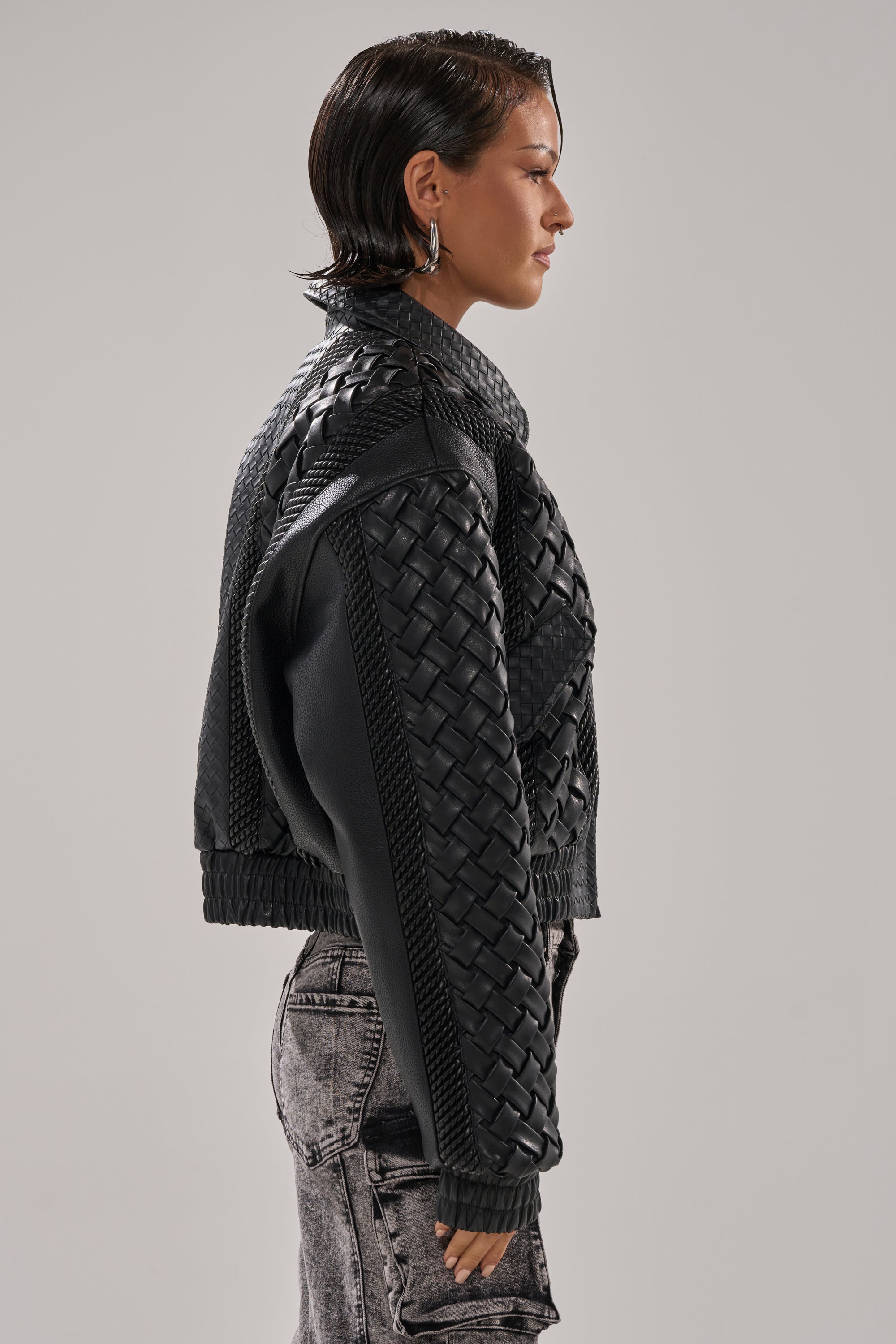 A person with short, wet-look hair is shown in profile wearing the KEEP IT LOWKEY PATCHWORK BOMBER and light gray acid-wash jeans against a plain gray background.