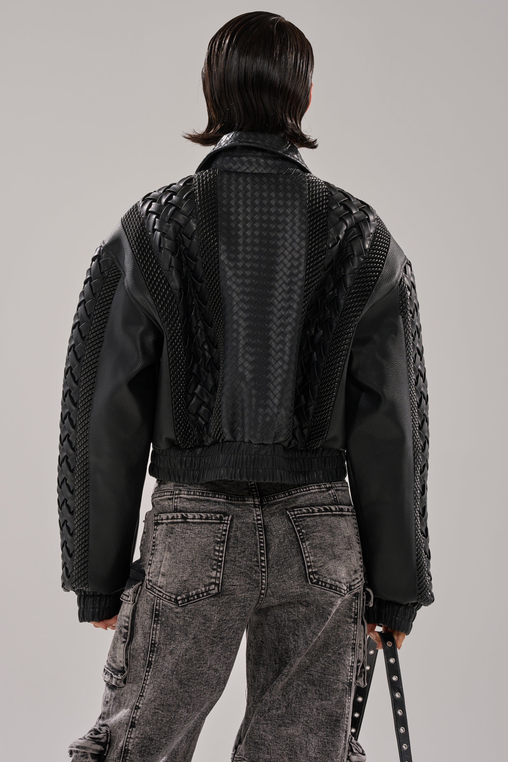 A person with straight, dark hair wears the KEEP IT LOWKEY PATCHWORK BOMBER—a black, textured leather jacket with woven accents—paired with gray acid-wash cargo jeans, standing facing away from the camera against a plain backdrop.