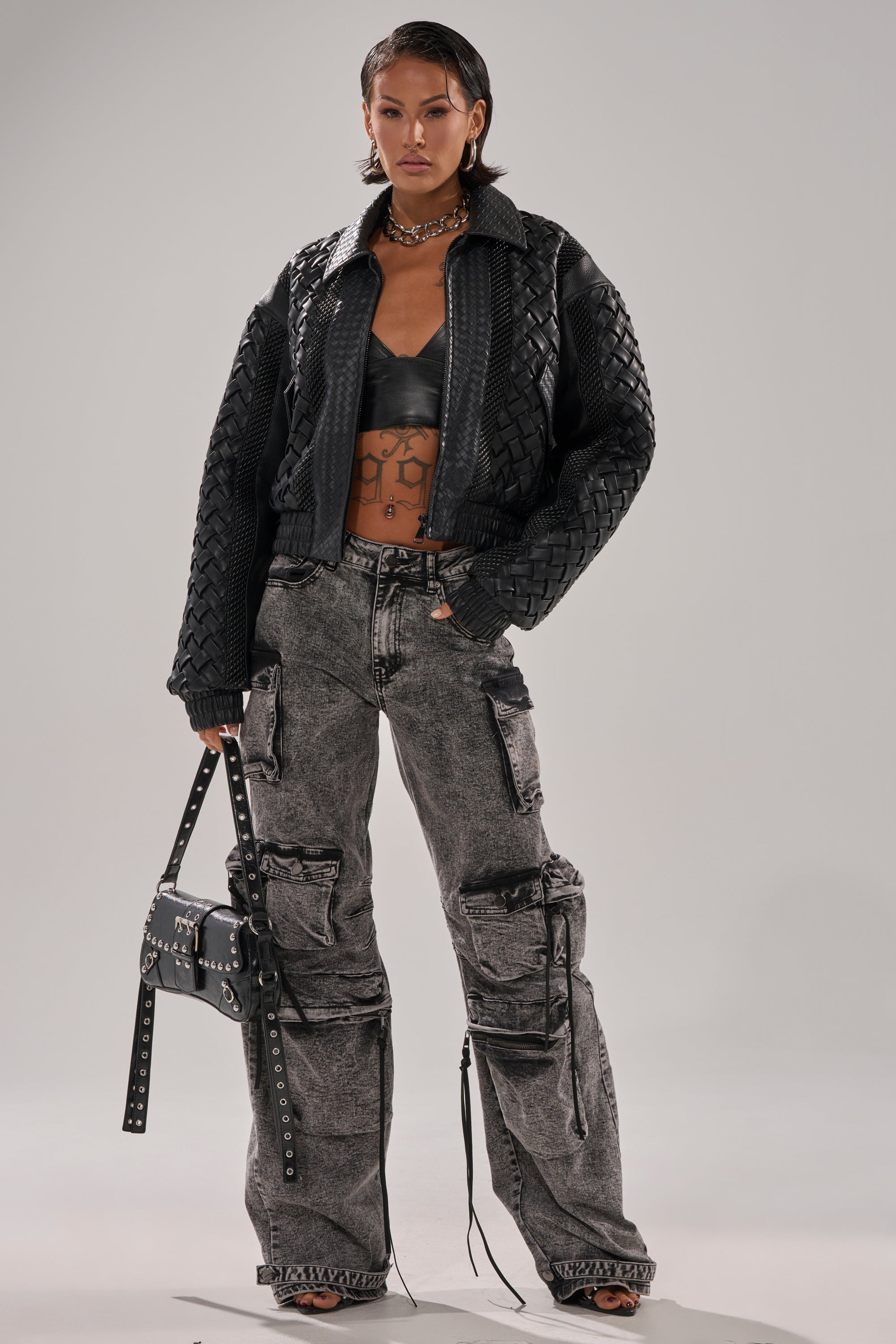 A person stands confidently in the KEEP IT LOWKEY PATCHWORK BOMBER, metallic bralette, gray acid-wash cargo pants, heels, and bold jewelry, holding a studded black handbag. They have short hair and pose against a plain backdrop.