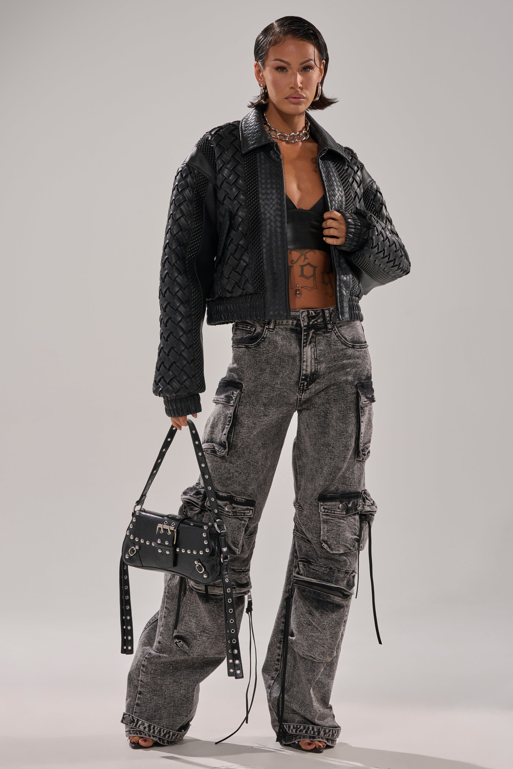 A woman poses in the KEEP IT LOWKEY PATCHWORK BOMBER, styled with a black bralette, gray cargo pants, studded black handbag, hoop earrings, and a choker against a plain background.