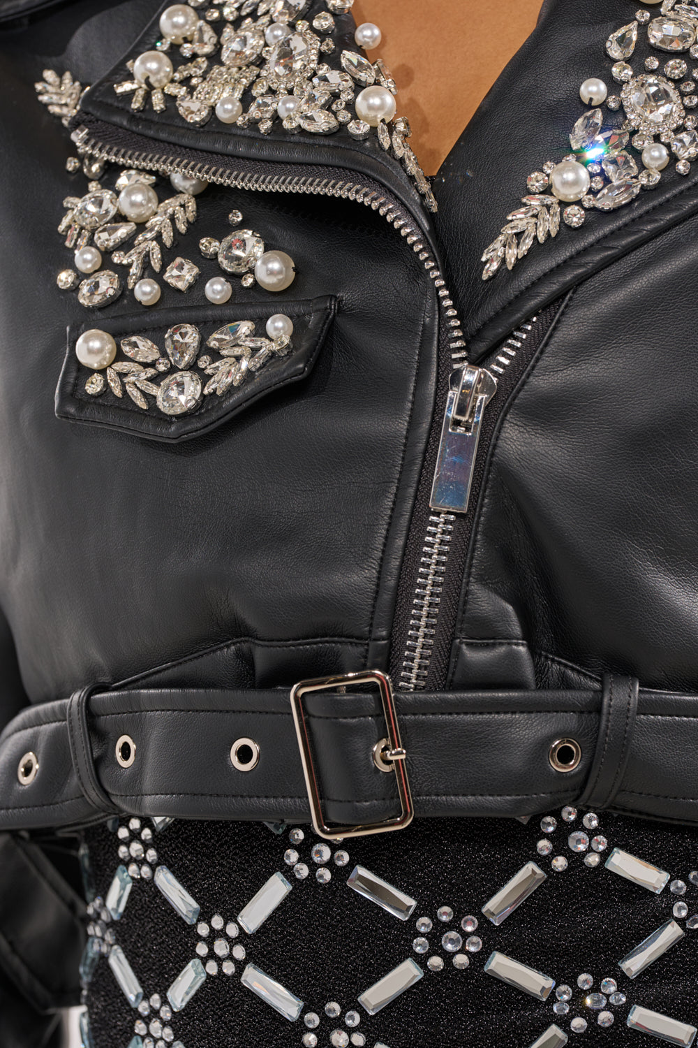 Close-up of the REINA EMBELLISHED MOTO faux leather jacket with silver zipper, belt, and floral pearl and rhinestone details, styled with a black skirt featuring silver geometric studs. 10