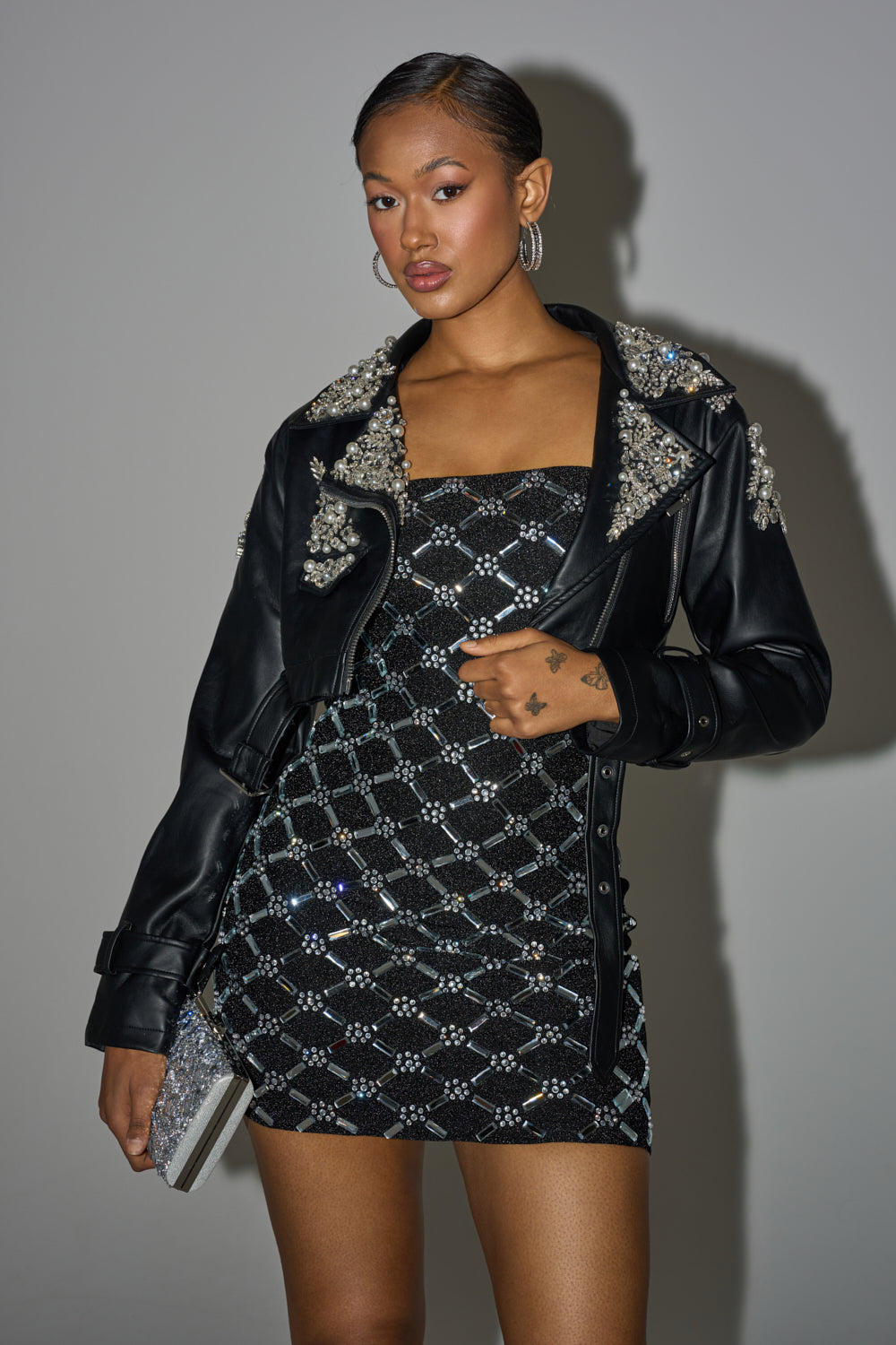 A woman stands confidently in the REINA EMBELLISHED MOTO jacket over a black embellished mini dress, holding a glittery clutch. She wears hoop earrings, has slicked-back hair, and shows off a hand tattoo. 3