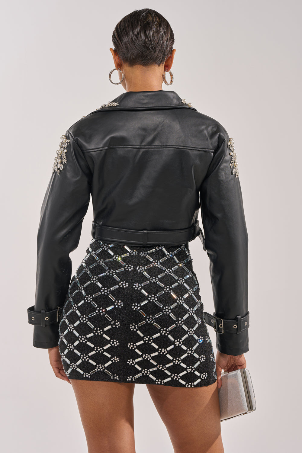 Seen from behind, a person with short hair wears the REINA EMBELLISHED MOTO faux leather cropped jacket with decorative shoulder details, a sparkly patterned mini skirt, and holds a small silver clutch. 6