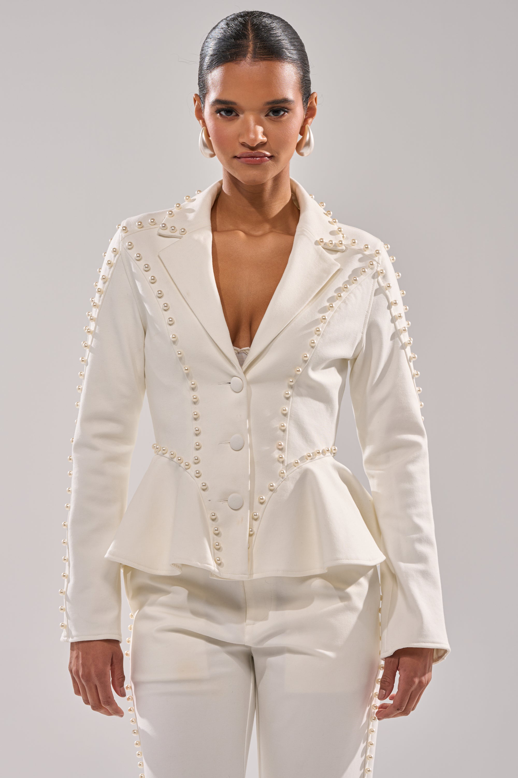 A woman stands facing forward, wearing the CHLOE PEARL BLAZER—a stylish white peplum blazer with pearl-like buttons—paired with matching white pants against a plain background. 0