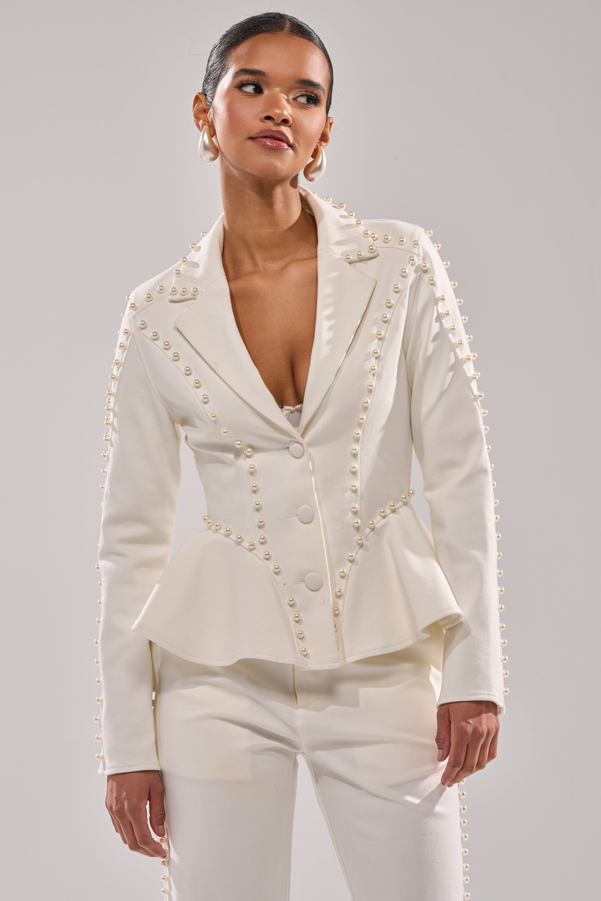 A woman models the CHLOE PEARL BLAZER—a sophisticated white blazer—styled with matching pants, set against a simple light gray background. 1
