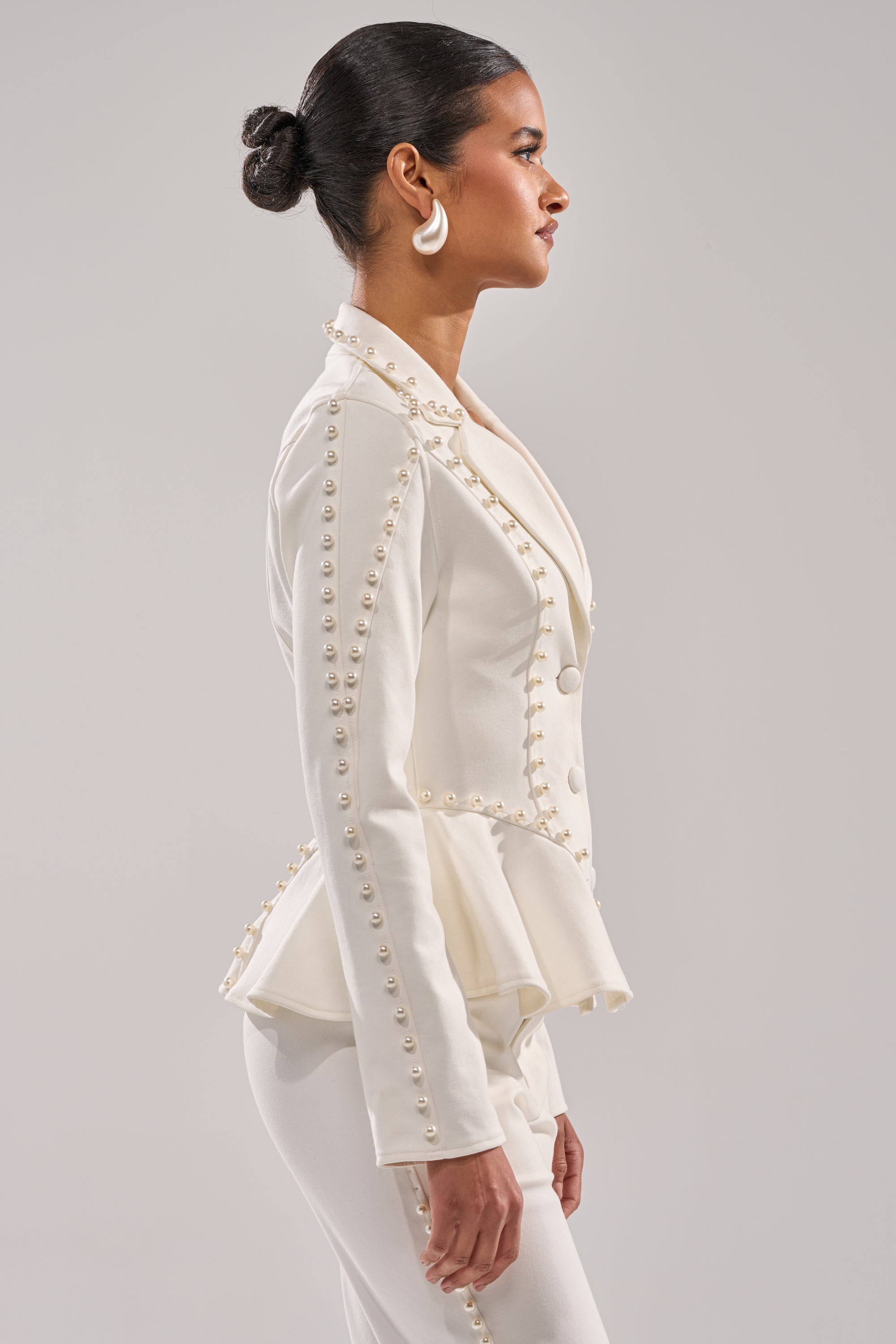 A woman in profile wears the CHLOE PEARL BLAZER, a fitted white peplum jacket with rows of pearl-like buttons and matching pants. Her hair is styled in a sleek bun, complemented by large white earrings. 3