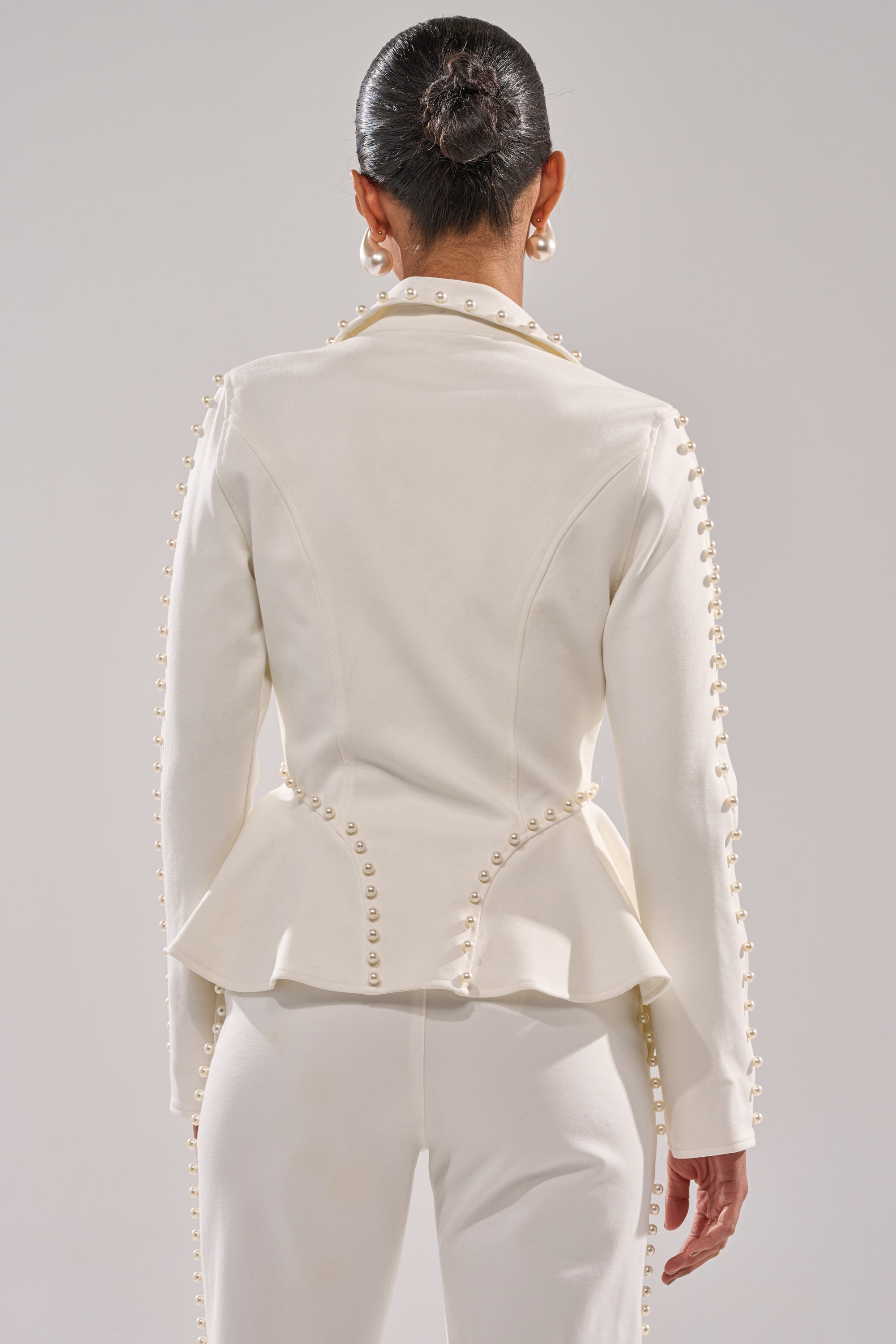 A person with dark hair in a neat bun is seen from behind, wearing the CHLOE PEARL BLAZER, distinguished by pearl embellishments on the seams and sleeves and elegant peplum detailing. 4