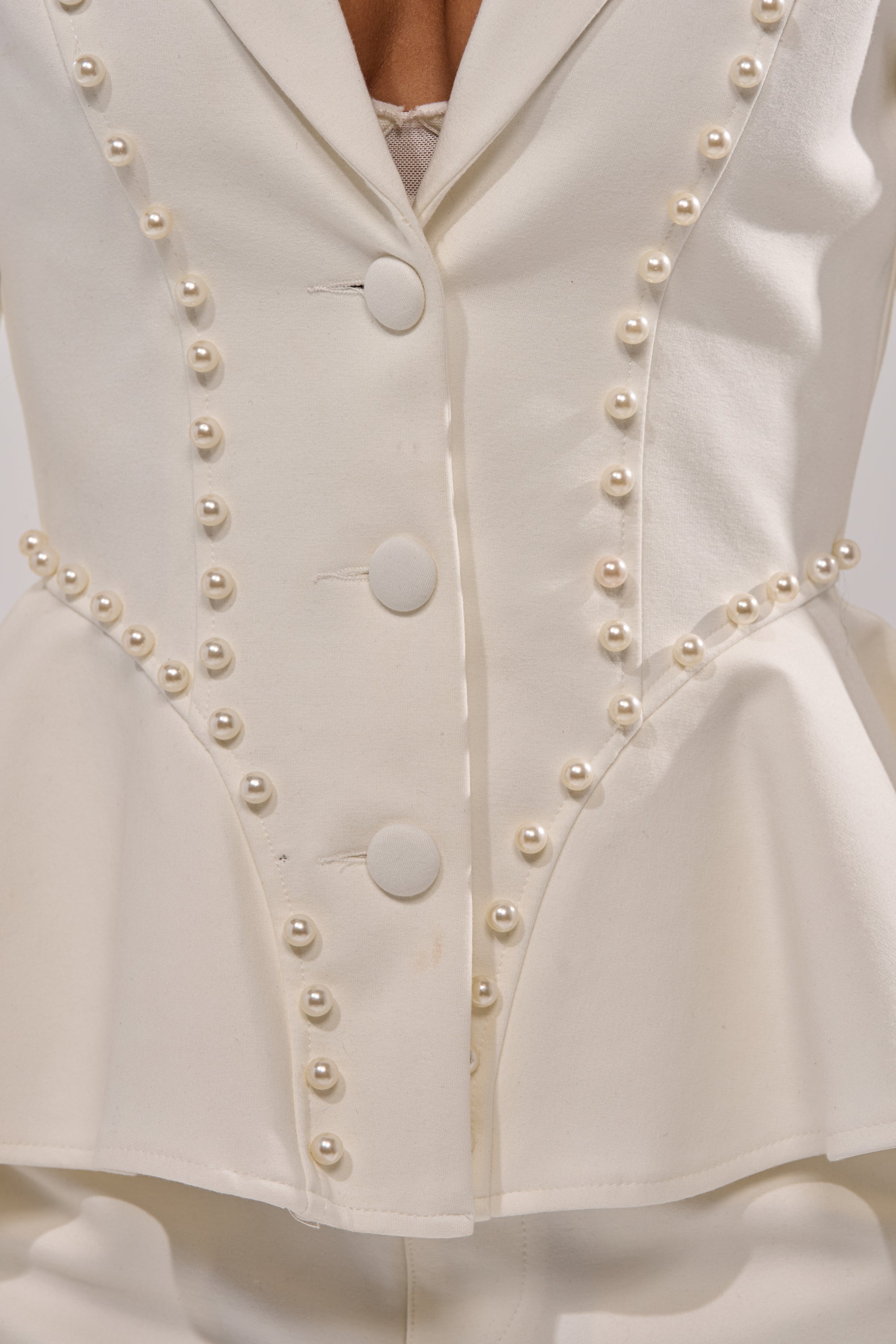 Close-up of the CHLOE PEARL BLAZER, a white tailored blazer featuring pearl embellishments, three fabric-covered buttons, and decorative pearls along the seams and waist, styled with matching white pants. 7