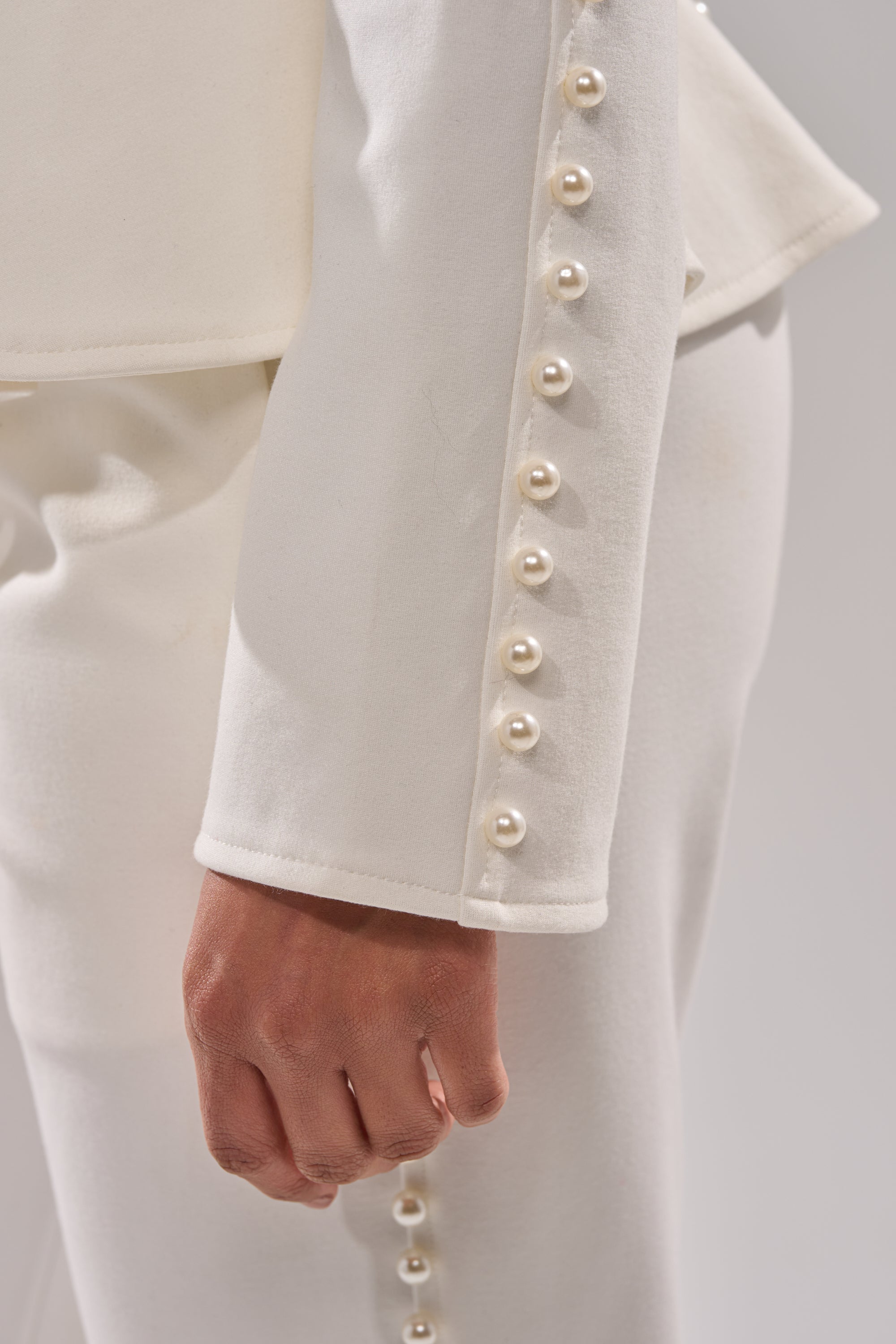 A close-up shows a person wearing the CHLOE PEARL BLAZER, featuring pearl buttons along the sleeve and pants. Their relaxed hand by their side highlights the luxurious pearl embellishments. 8