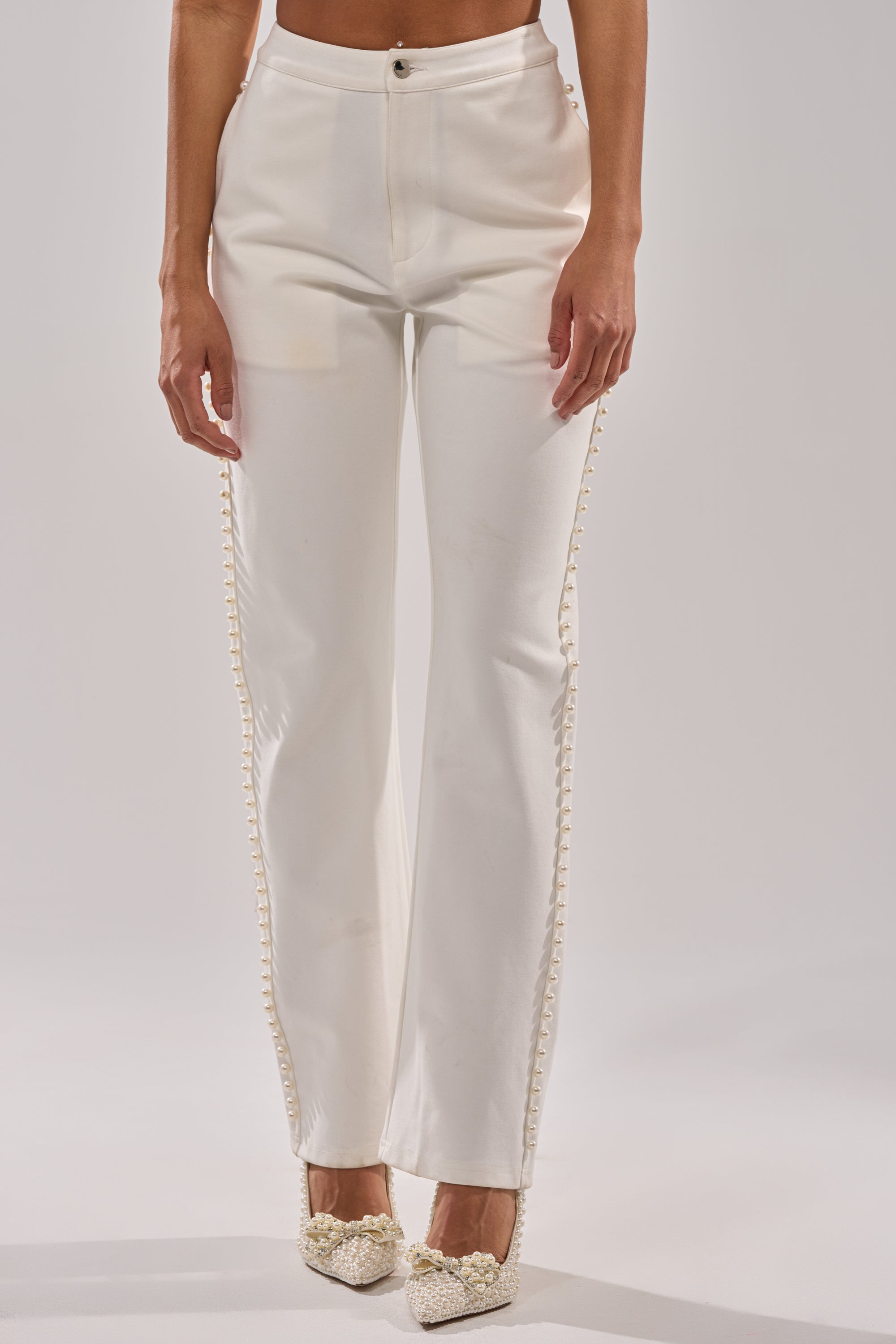 Wearing the CHLOE PEARL TROUSER, which features high-rise cotton fabric and decorative side buttons, paired with white embellished shoes, a person stands against a plain light background. 0