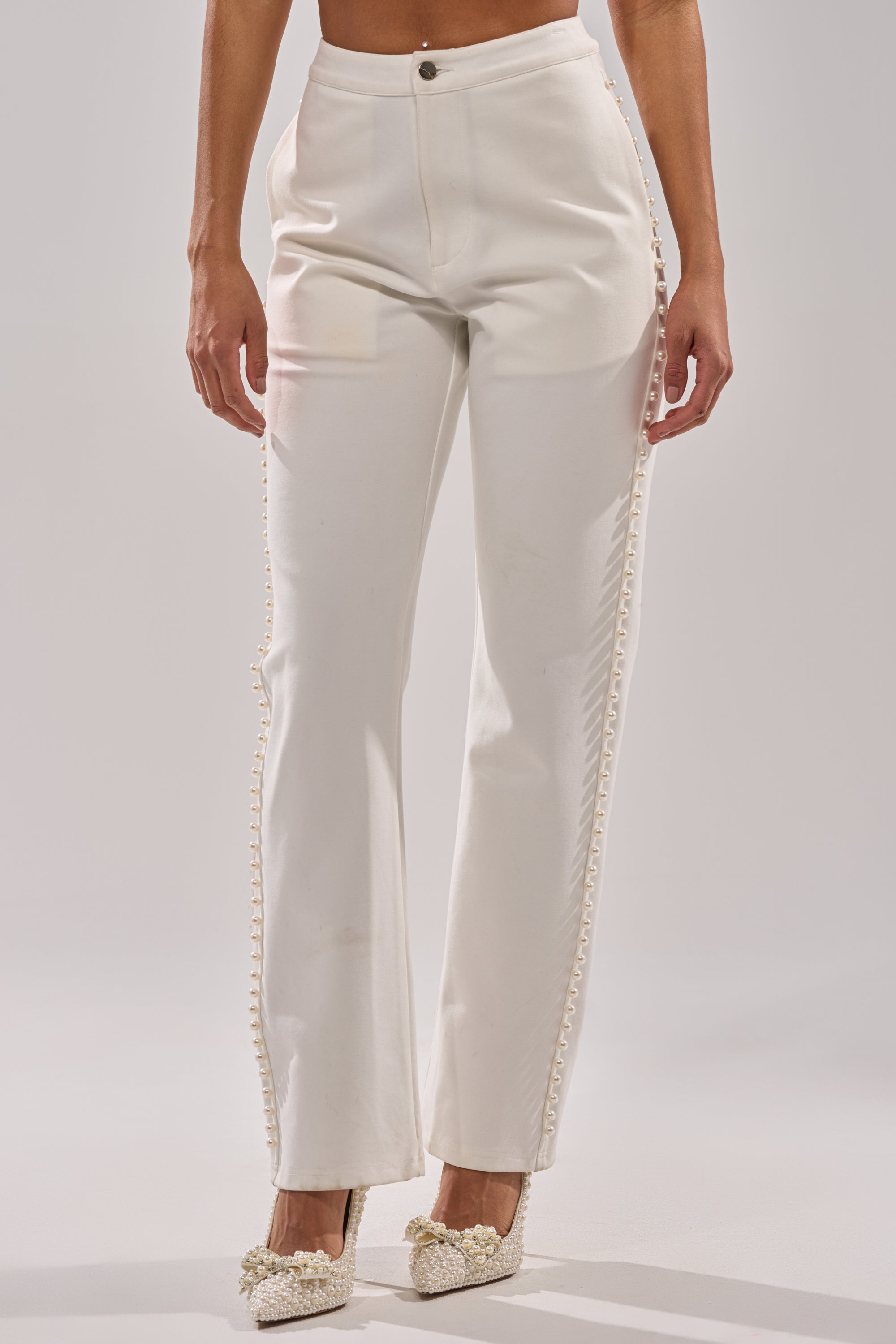 A person stands against a plain light background wearing the CHLOE PEARL TROUSER, high-rise white pants with decorative side trim, paired with off-white embellished heels featuring faux pearls. 1