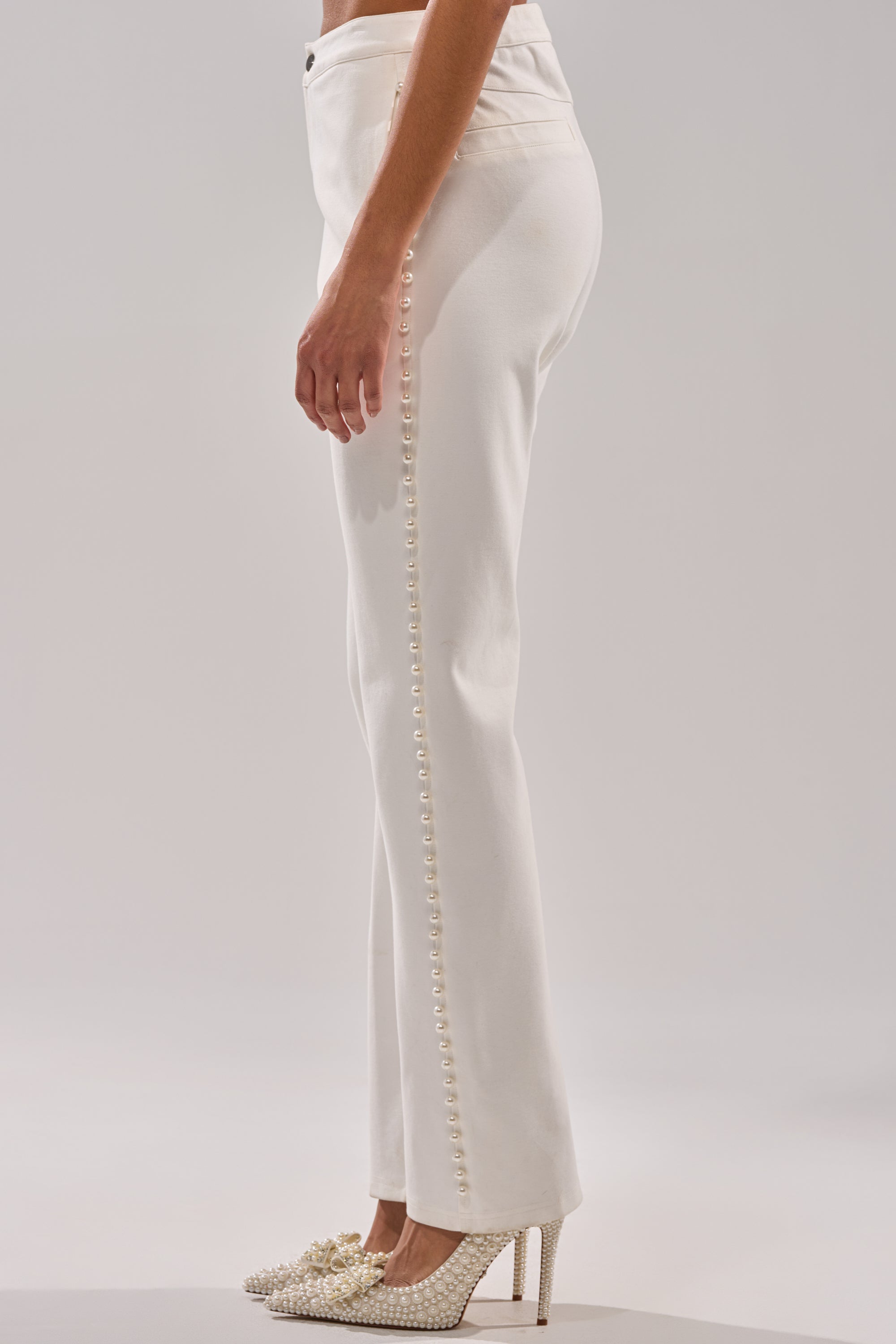 A person models the CHLOE PEARL TROUSER—high-rise white pants with faux pearl details on the side seam—paired with pearl-adorned high heels, set against a plain, light-colored background. 2