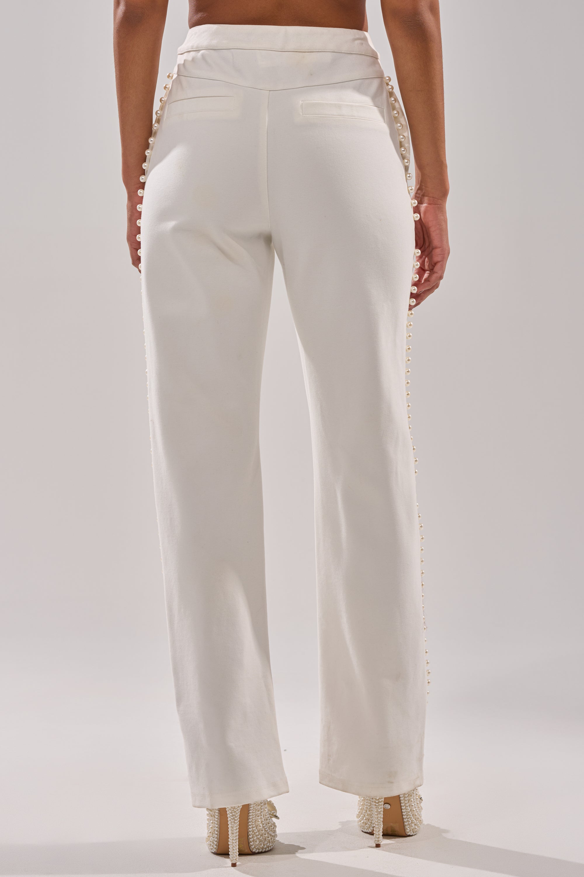 A person is photographed from the back wearing the CHLOE PEARL TROUSER, a high-rise, white straight-leg pant with decorative pearl-like buttons along the side seams and welt pockets. 3
