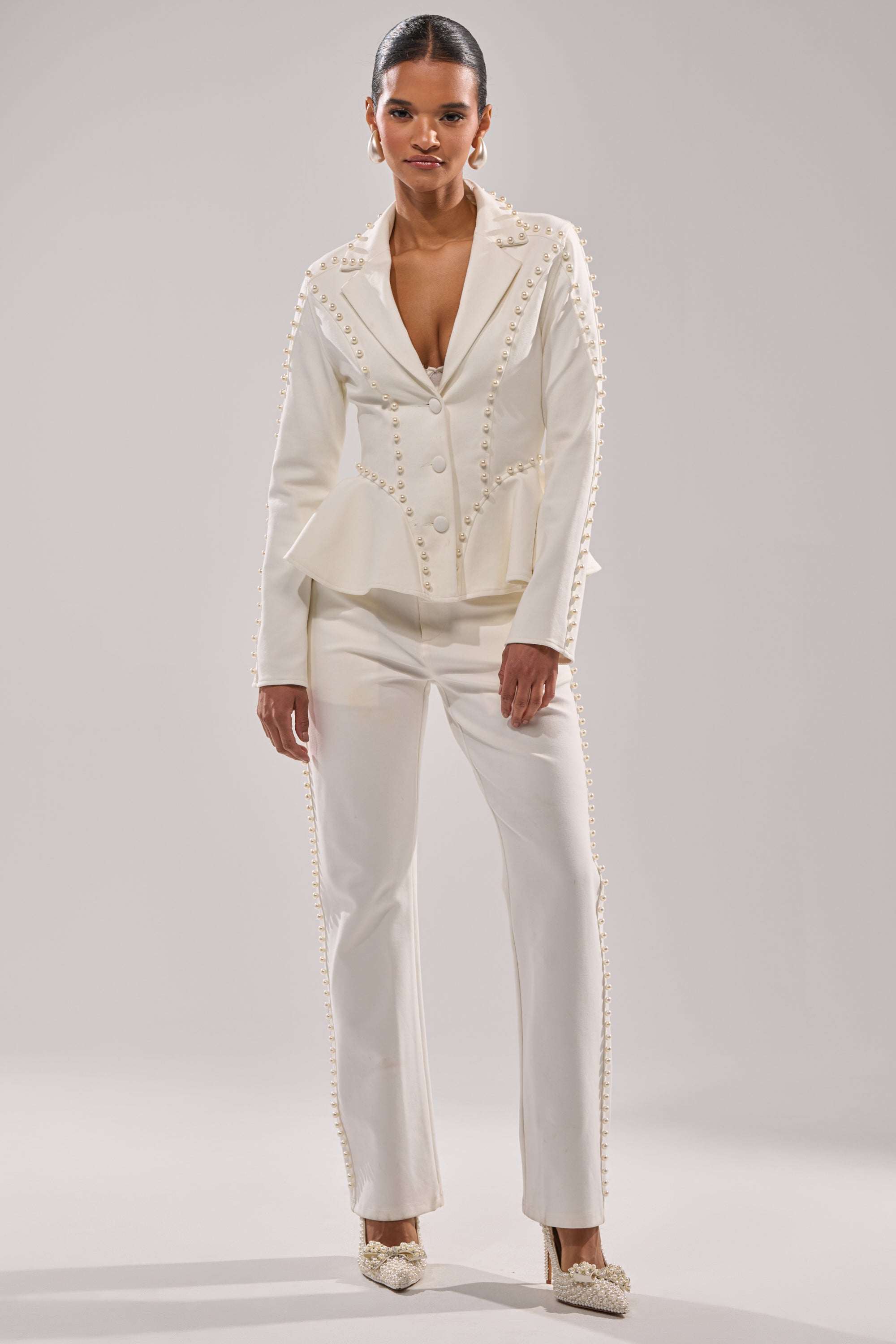 A woman stands facing forward in the CHLOE PEARL TROUSER, a tailored white pantsuit embellished with pearls and paired with white heels, set against a plain light gray background. 4