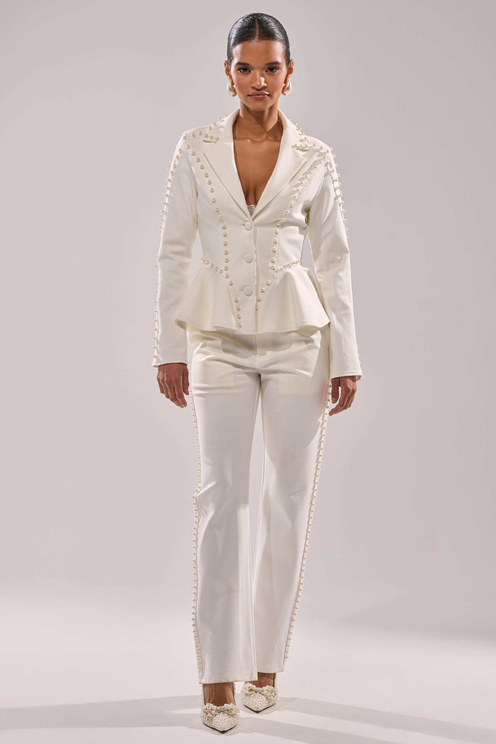 A woman stands confidently in the CHLOE PEARL TROUSER—a tailored white pantsuit featuring pearl-embellished pants and decorative accents—paired with pointed white heels, set against a plain light background. 5