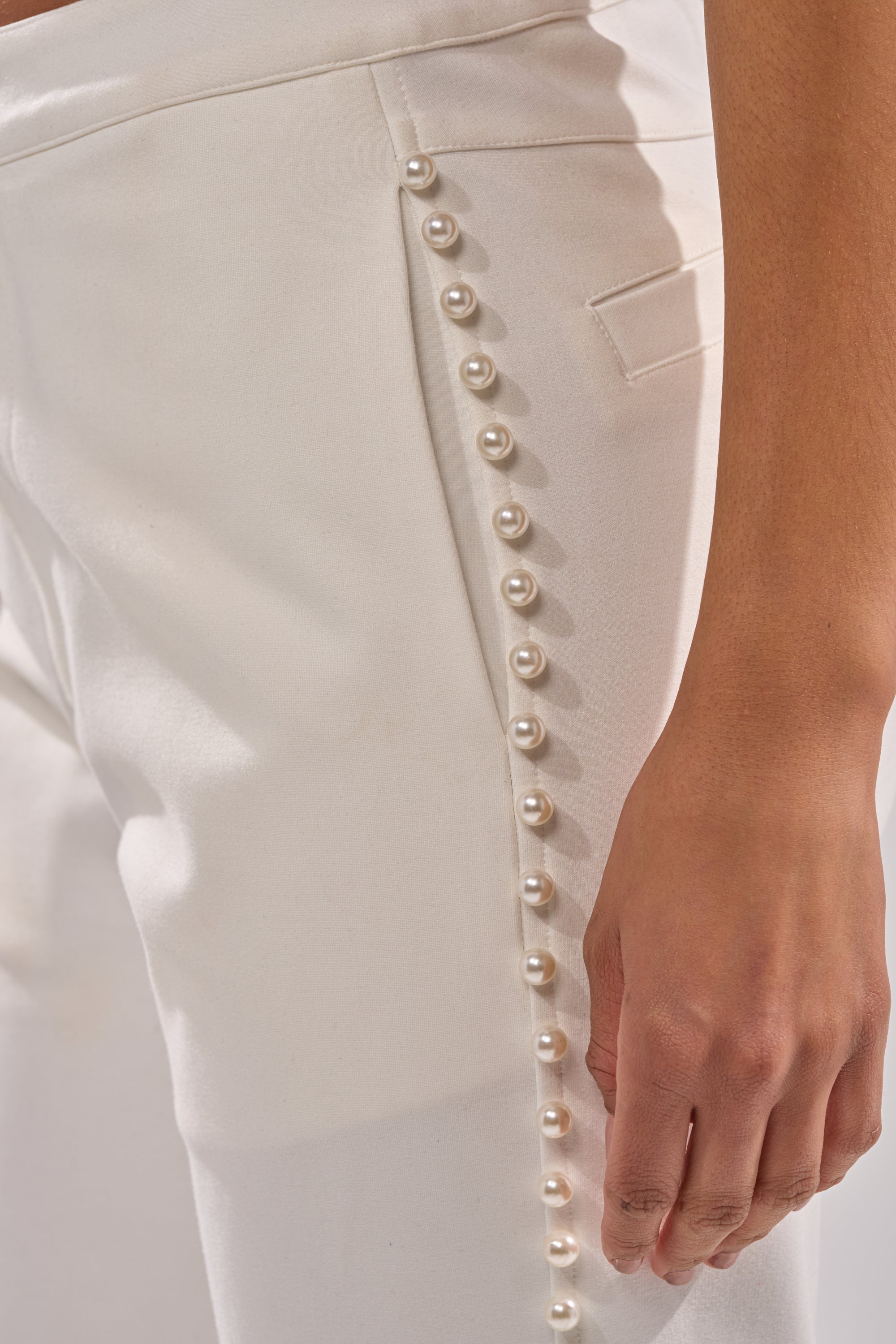 Close-up of the CHLOE PEARL TROUSER, high-rise cotton pants with a row of decorative pearl buttons along the side seam from waist to hip. A hand rests beside these elegant, pearl-embellished trousers near the thigh. 6