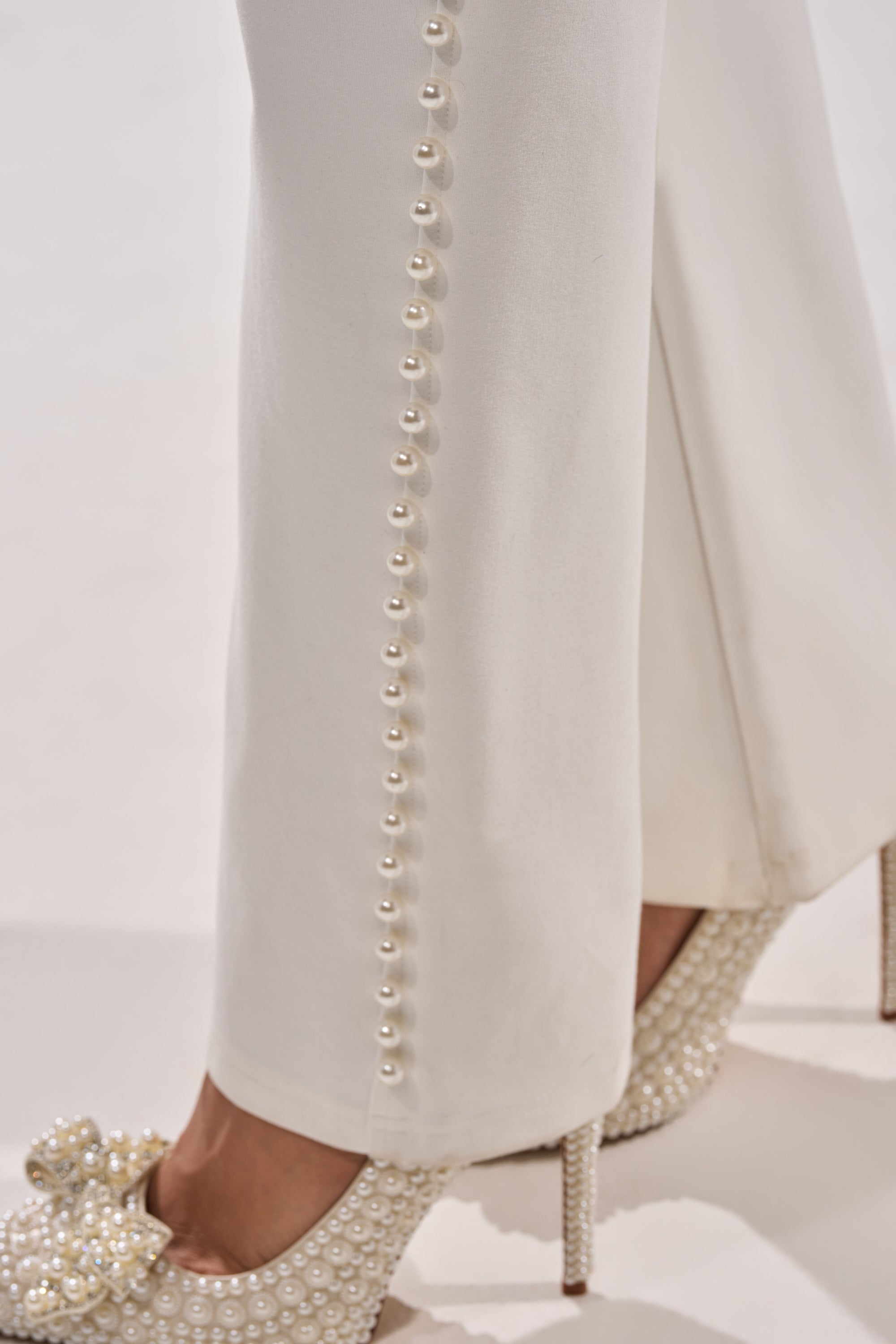 Close-up of a person in CHLOE PEARL TROUSER, featuring pearls along the side seam, paired with white heels embellished with pearls and a beaded bow, standing on a light background. 7