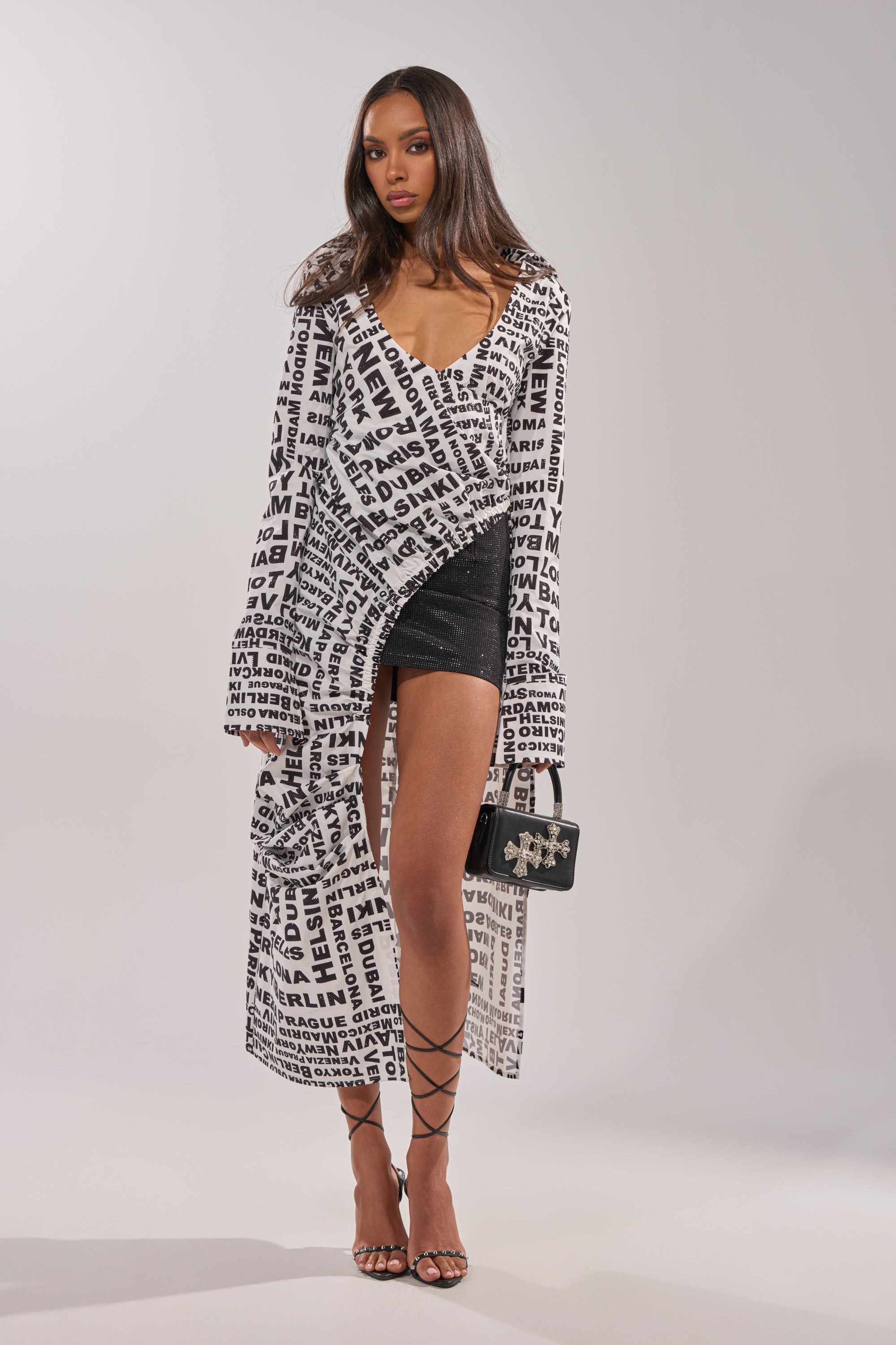 A woman wears the ALL AROUND THE WORLD BLOUSE—a long, white top with typography print—over a sparkly black dress. She accessorizes with a small black purse, strappy high-heeled sandals, and has straight, loose hair. 0
