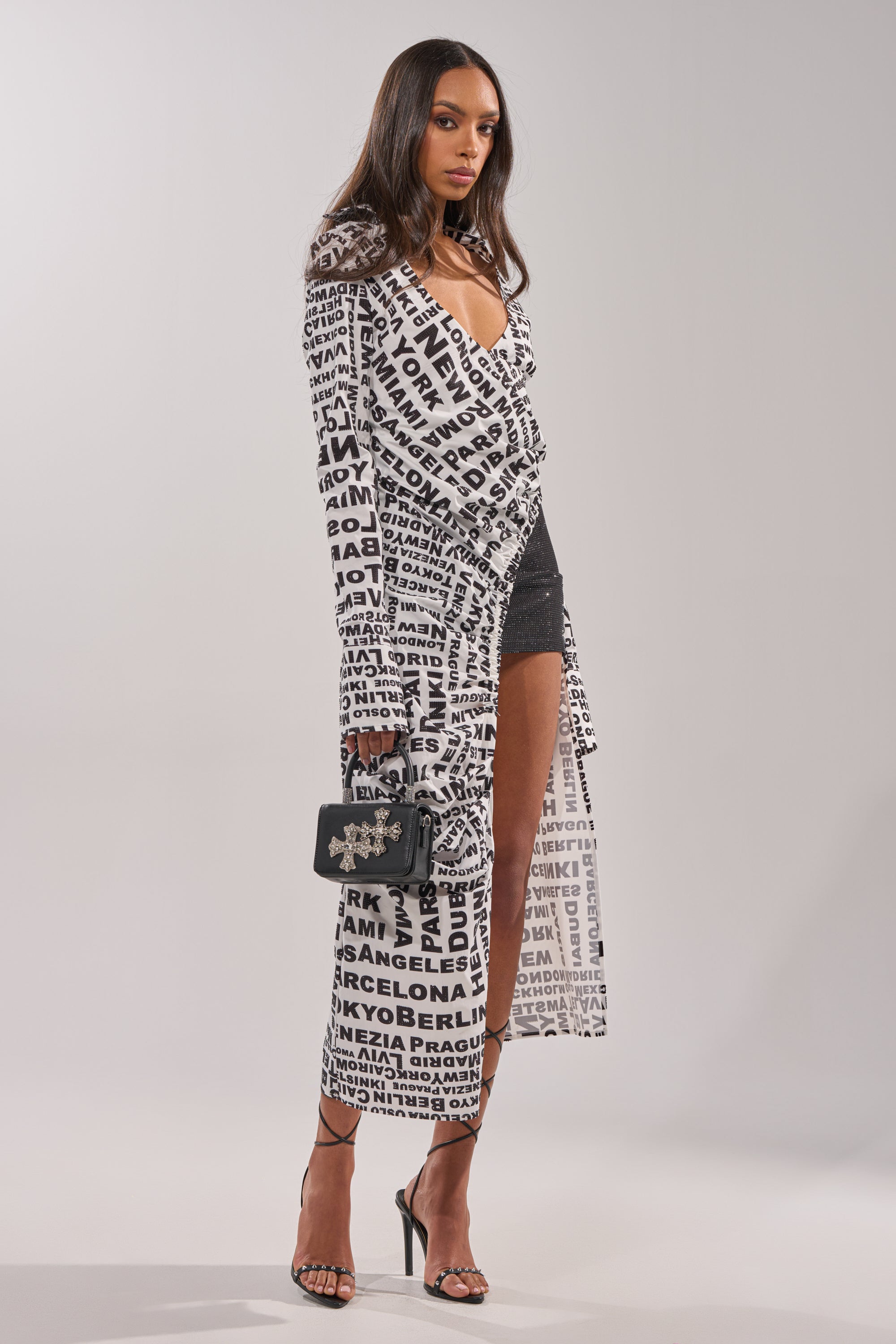 A woman models the ALL AROUND THE WORLD BLOUSE—a floor-length, collared blouse with black city names—over a black mini dress, styled with high heels and a small black handbag against a plain light background. 3