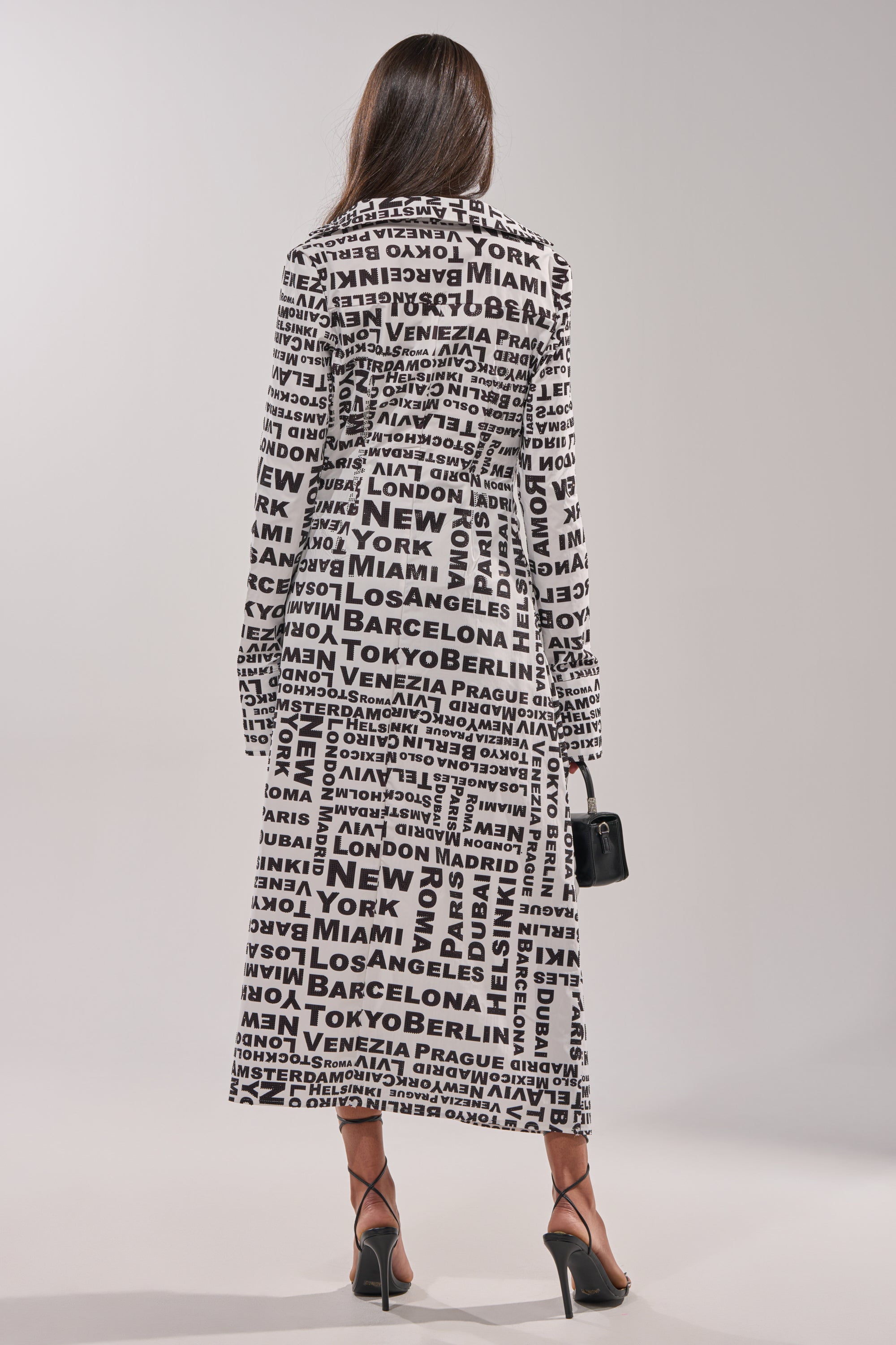 A woman faces away, wearing the ALL AROUND THE WORLD BLOUSE—a statement black-and-white, city-print wrap styled as a long coat. She carries a small black handbag and wears black strappy heels against a plain background. 6