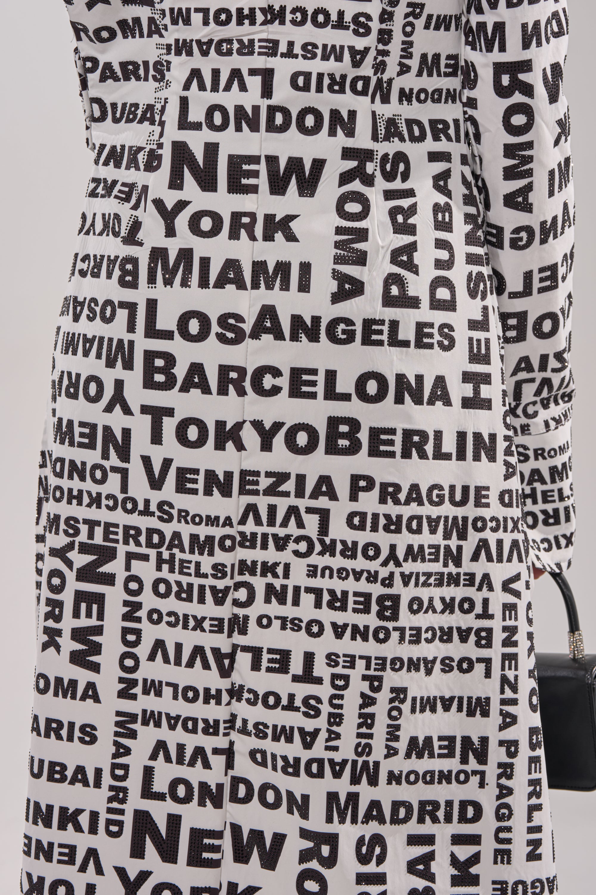 A close-up of a person wearing the ALL AROUND THE WORLD BLOUSE, a floor-length collared blouse featuring bold black city names like New York, Tokyo, and Berlin in various sizes and orientations. 8