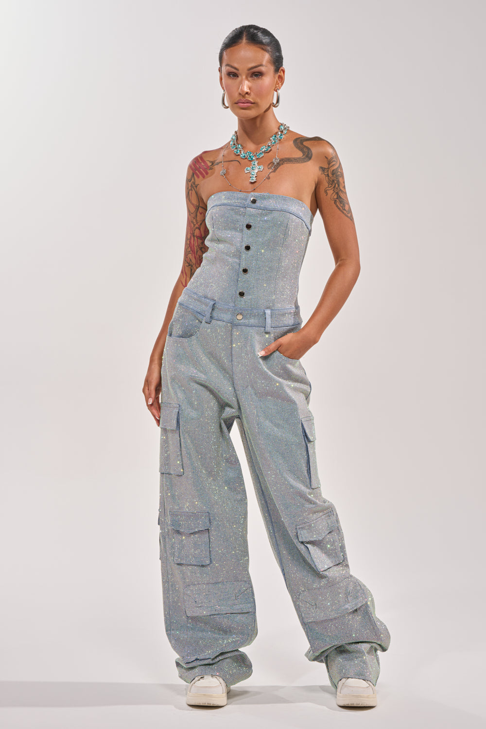 A woman exudes confidence in the TOO FLY RHINESTONE DENIM JUMPSUIT, featuring cargo pockets and button details. She accessorizes with bold jewelry, visible tattoos, and white sneakers against a simple light background. 2